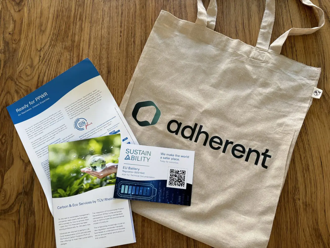 A canvas tote bag with "adherent" logo, a blue and white brochure on EU packaging regulations, and a white card about EU Battery Regulation with a QR code, all on a wooden table.