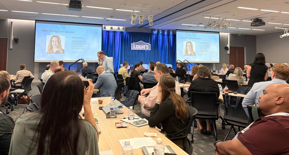 A conference room filled with people seated at tables, facing two large screens displaying 'Meet Our Keynote Speaker Monica Welt' and the Lowe's logo on a blue curtain backdrop.