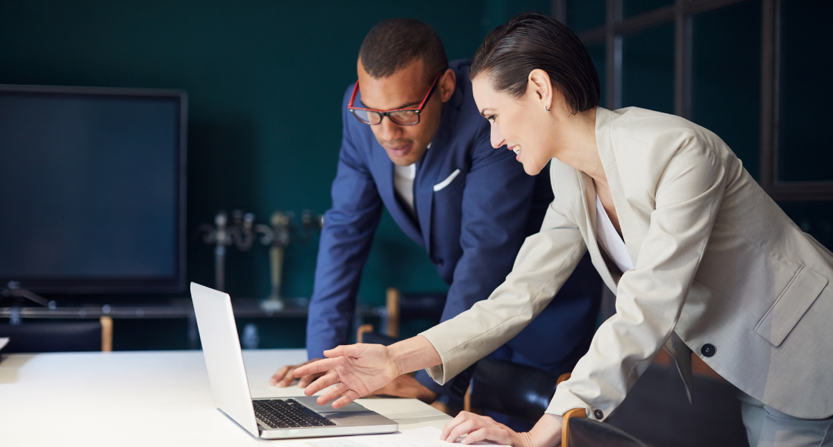 Two professionals, a man and a woman, lean over a table, looking at a laptop. The woman points at the screen.