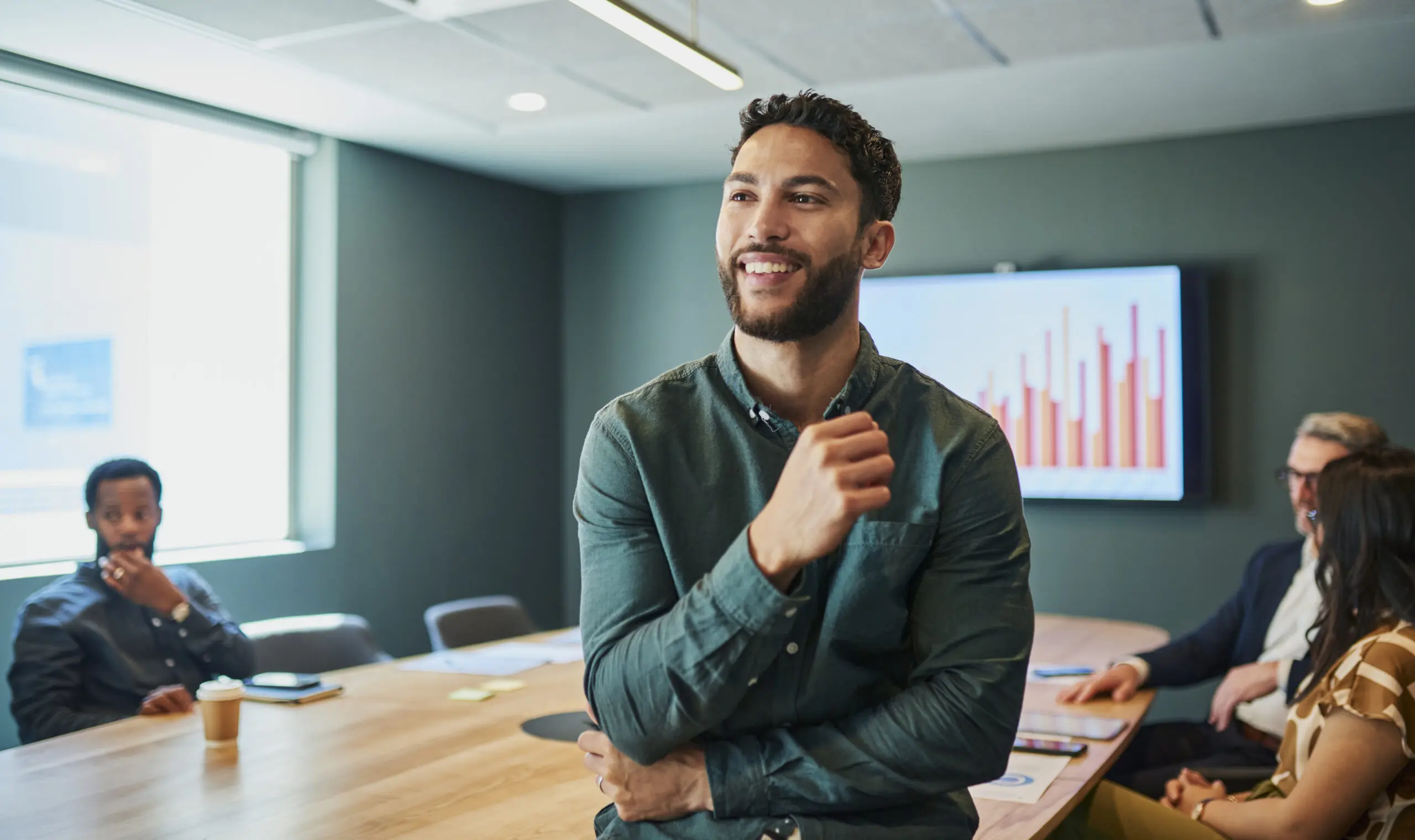 Adherent (formerly Compliance & Risks). A smiling man in a green shirt stands in a conference room, looking left. Colleagues are seated at a table behind him, and a screen displays a bar chart.