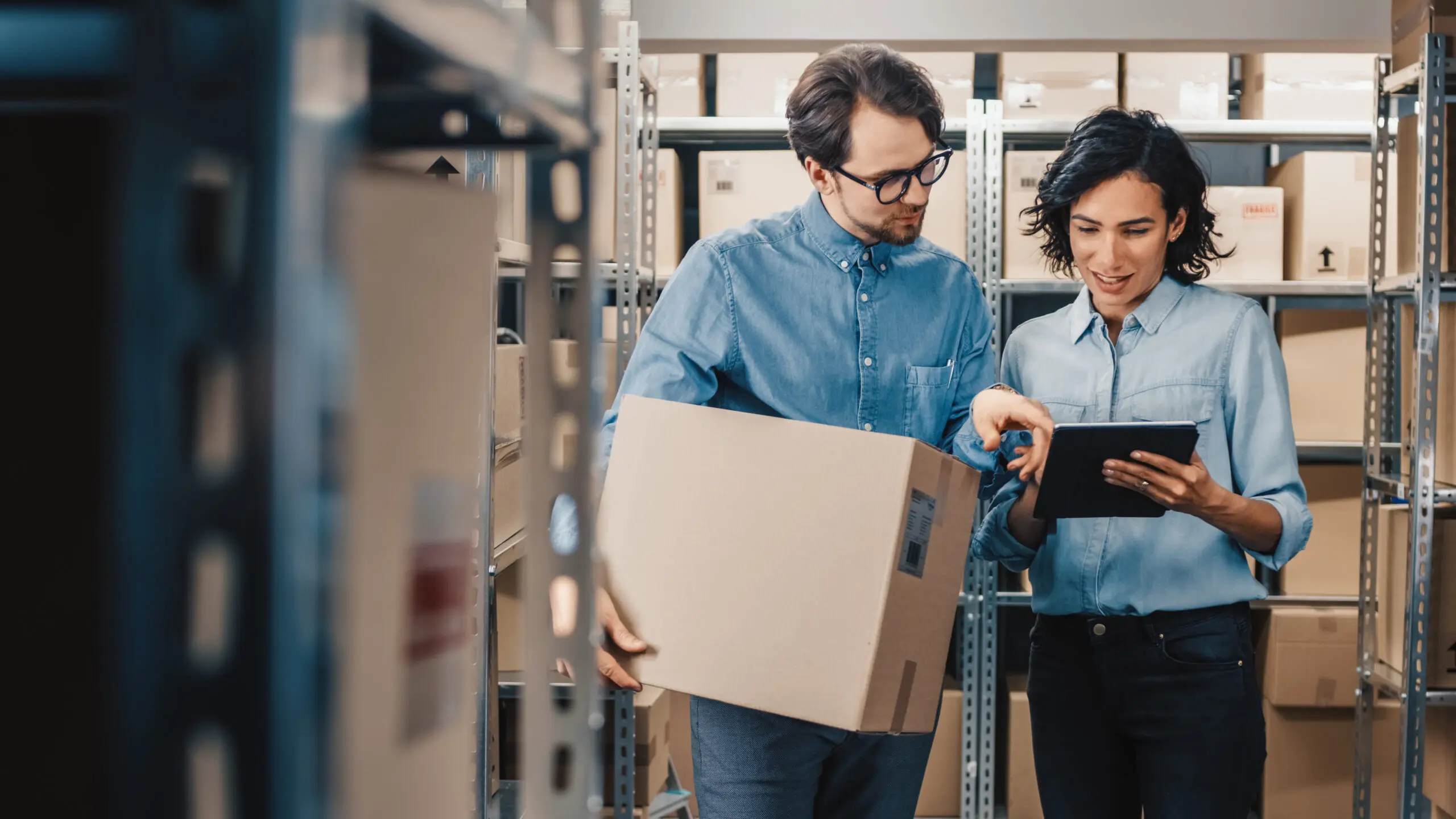 Adherent (formerly Compliance & Risks). A man holding a cardboard box and a woman holding a tablet look at the screen together in a warehouse filled with shelves of boxes.