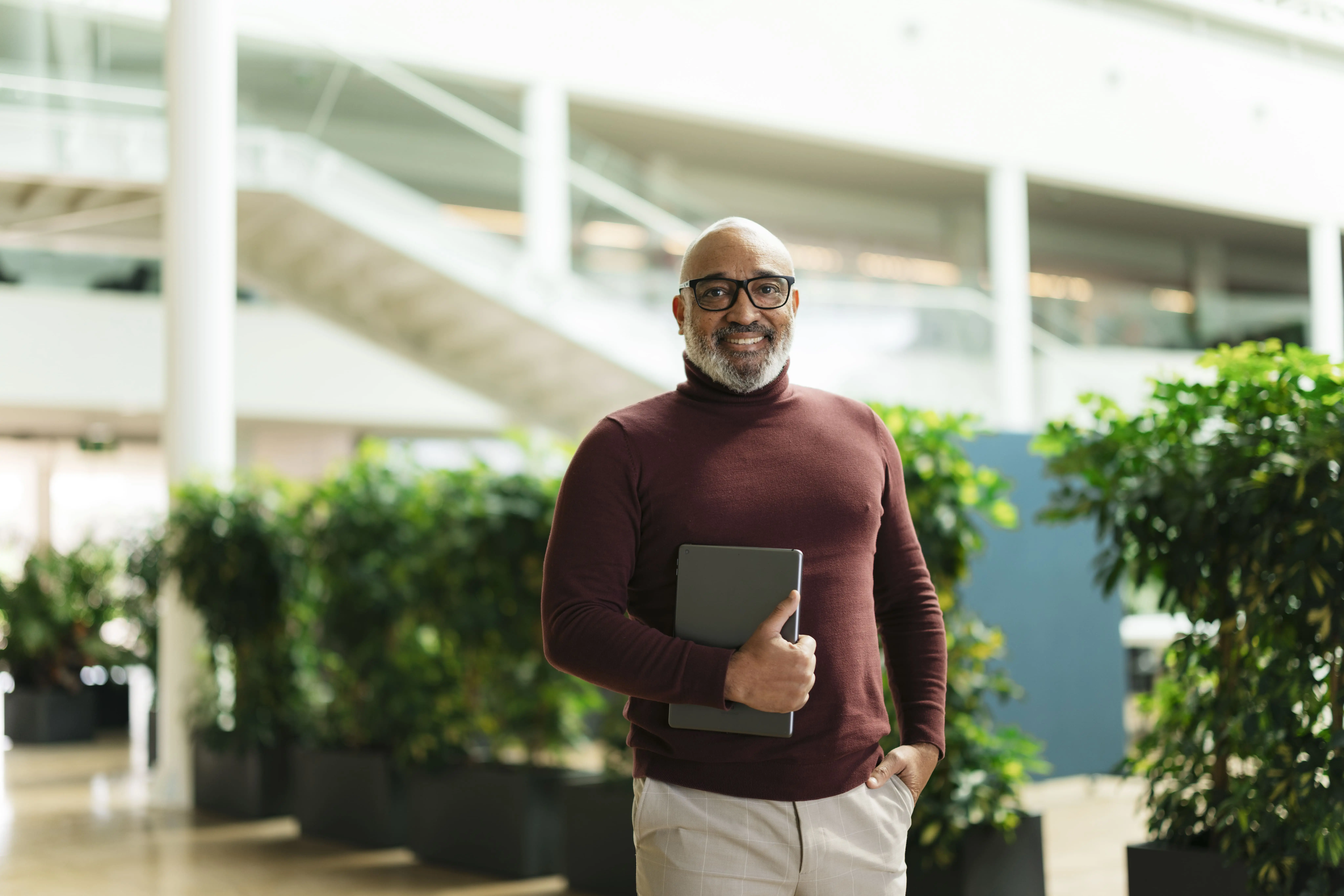 Adherent (formerly Compliance & Risks). A smiling man with a white beard and glasses, wearing a maroon turtleneck, holds a tablet in a modern office building.