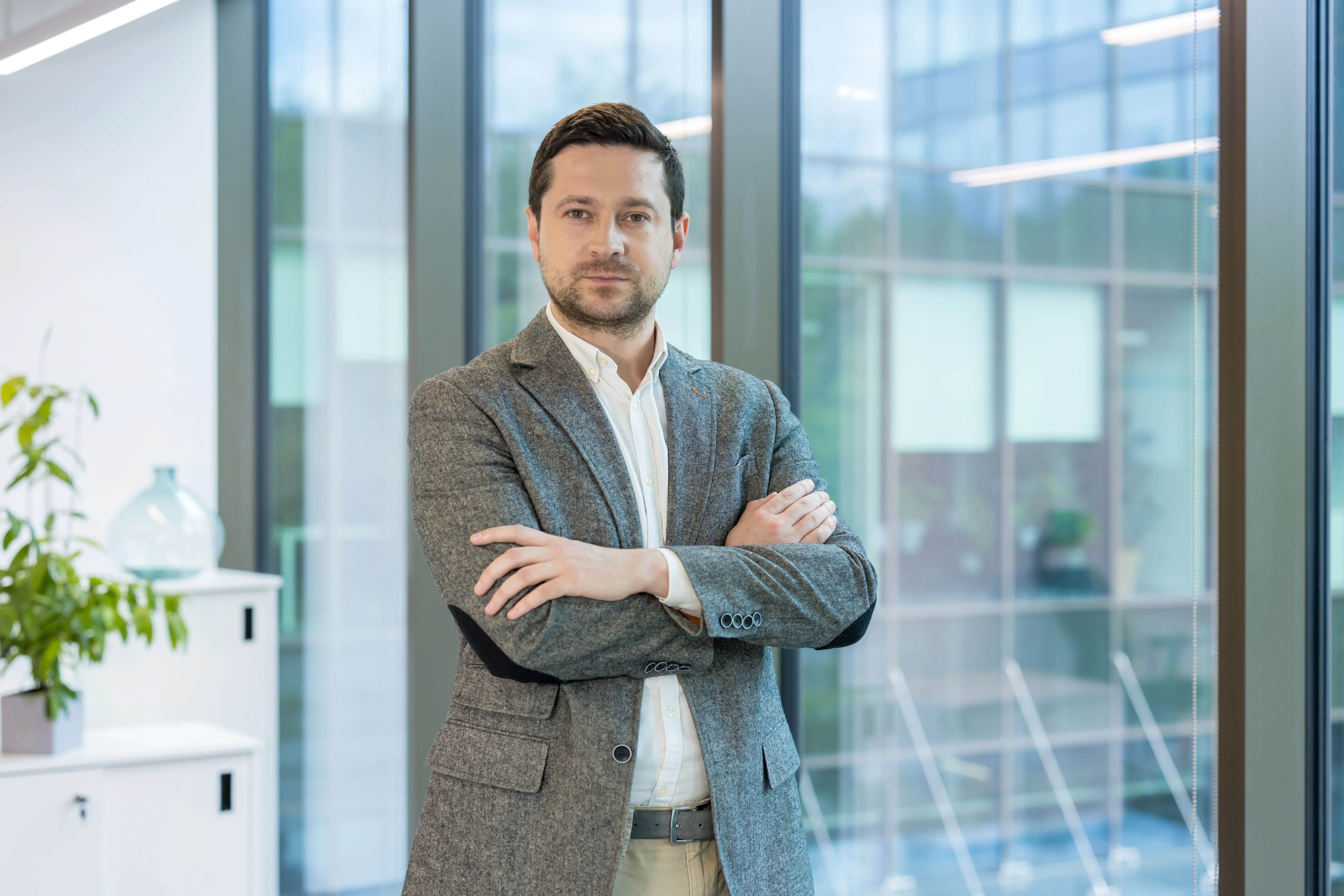 Adherent (formerly Compliance & Risks). A man in a grey blazer and white shirt stands with crossed arms in a bright office with large windows.