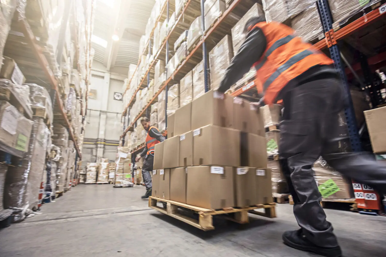 Adherent (formerly Compliance & Risks). Two warehouse workers in orange vests moving stacks of cardboard boxes on a pallet jack amidst tall shelving units filled with inventory, conveying a sense of busy operations.