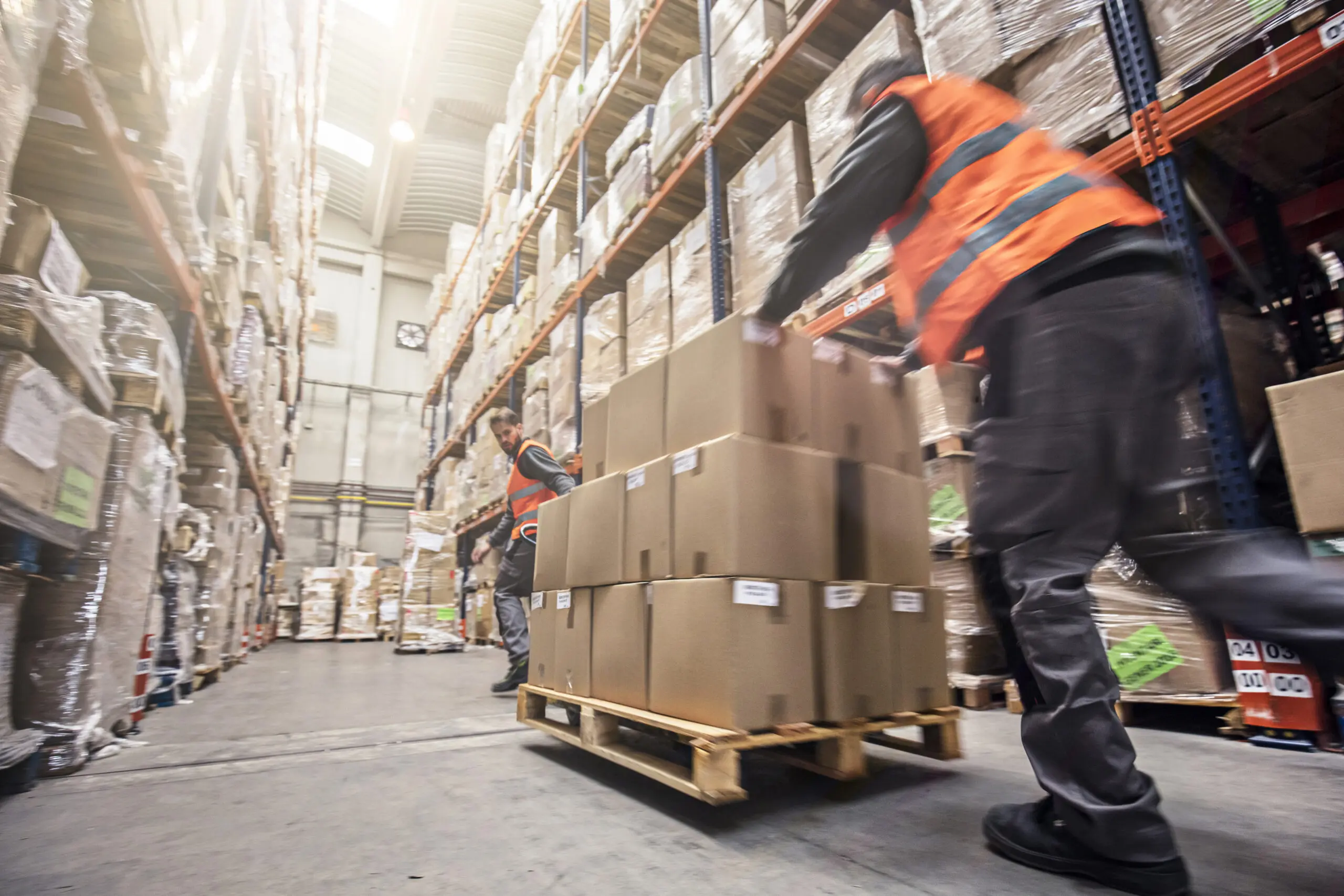 Two warehouse workers in orange vests moving stacks of cardboard boxes on a pallet jack amidst tall shelving units filled with inventory, conveying a sense of busy operations.
