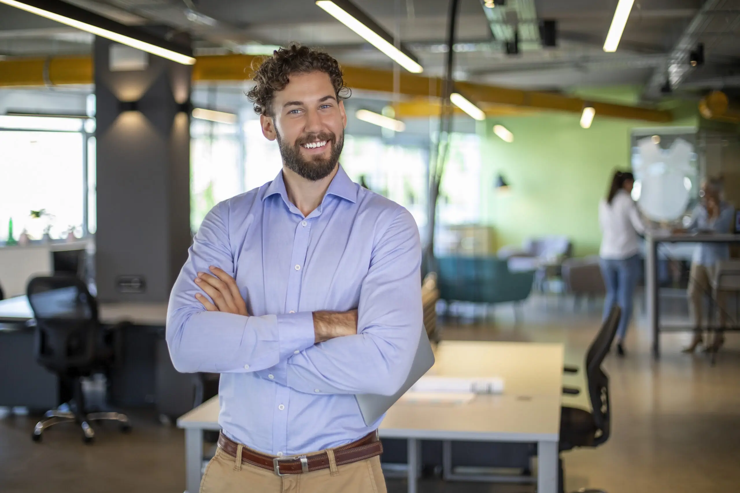 Adherent (formerly Compliance & Risks). A smiling man with curly hair and a beard, wearing a blue shirt and khaki pants, stands with crossed arms holding a laptop in a modern office.