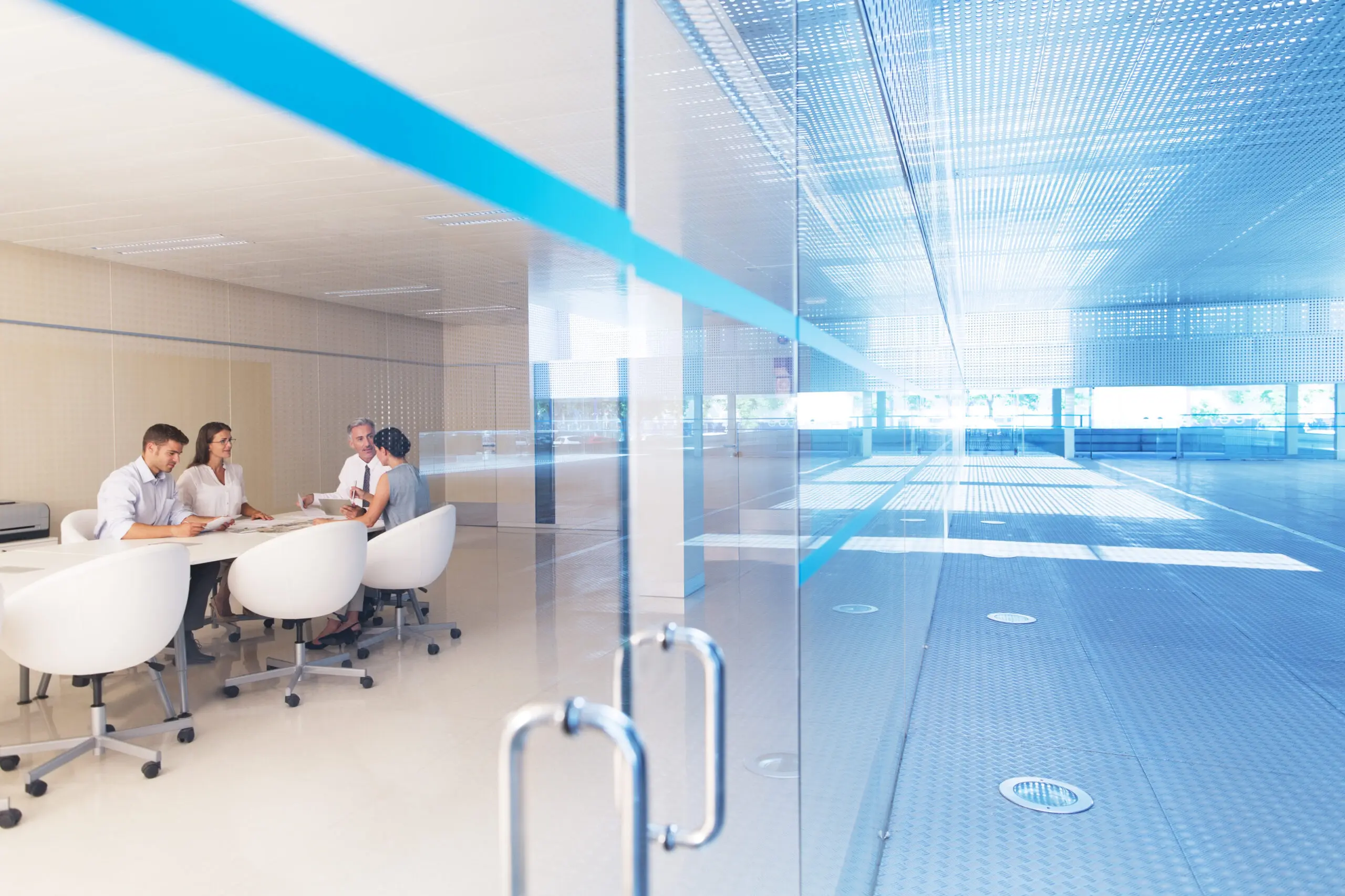Adherent (formerly Compliance & Risks). Four professionals in a meeting room, seen through a glass partition with a blue stripe, next to a bright, empty blue-floored open office area.