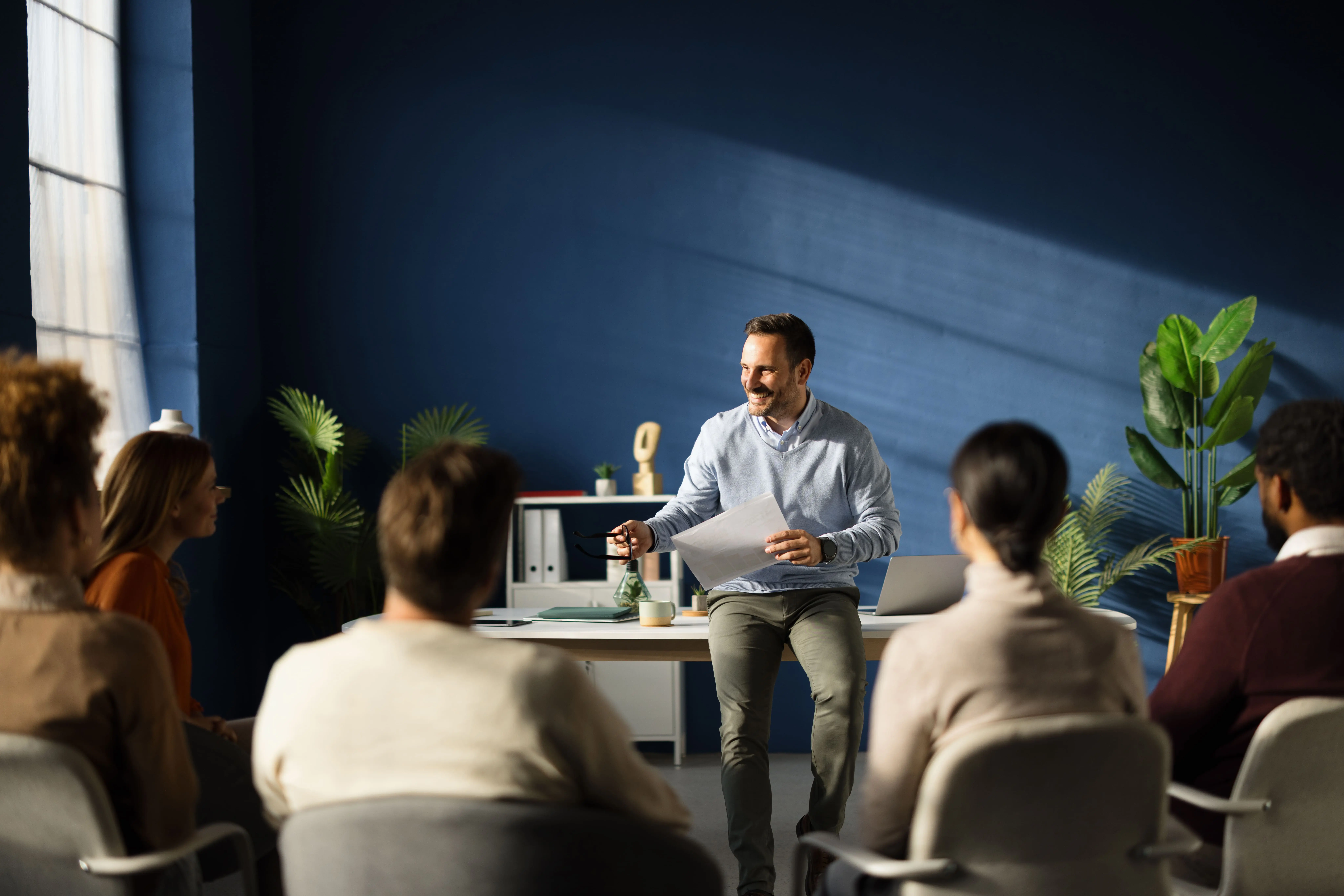 Adherent (formerly Compliance & Risks). A man in a blue sweater sits on a white desk, holding papers and glasses, smiling at a group of people seated in front of him in a modern office with a dark blue wall and large window.