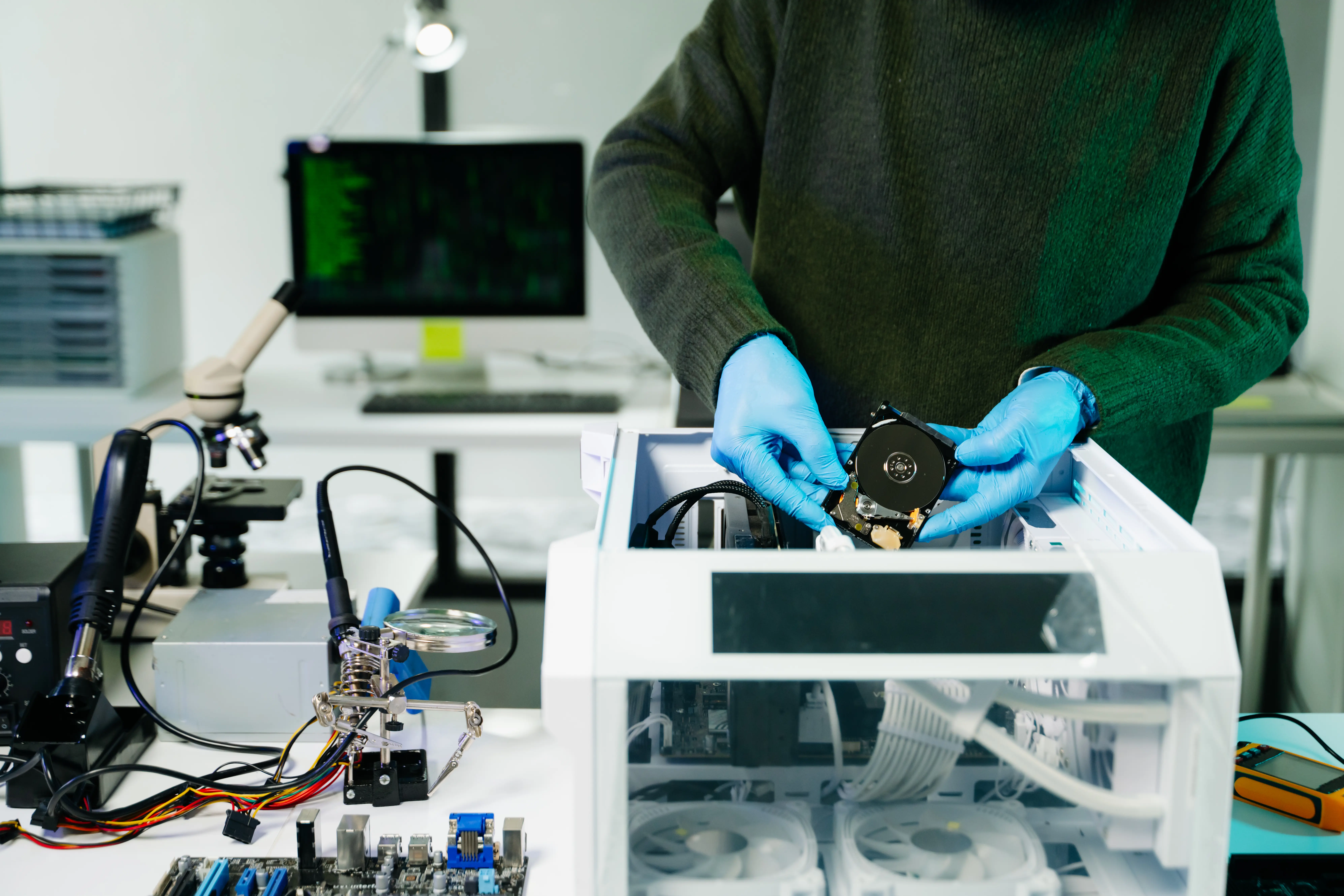 Adherent (formerly Compliance & Risks). Person in blue gloves installing a hard drive into a white computer case, surrounded by electronic tools and components.