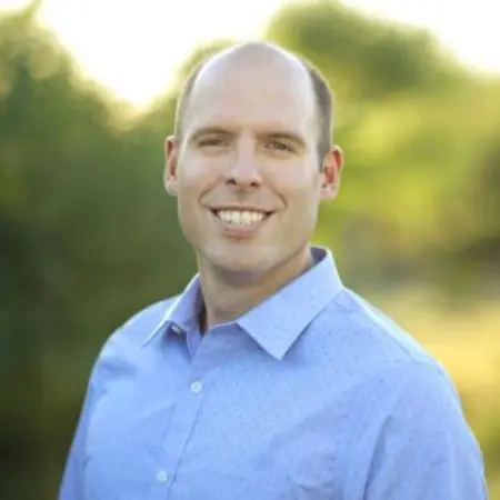 Headshot of a smiling man with a bald head wearing a light blue button-down shirt outdoors.