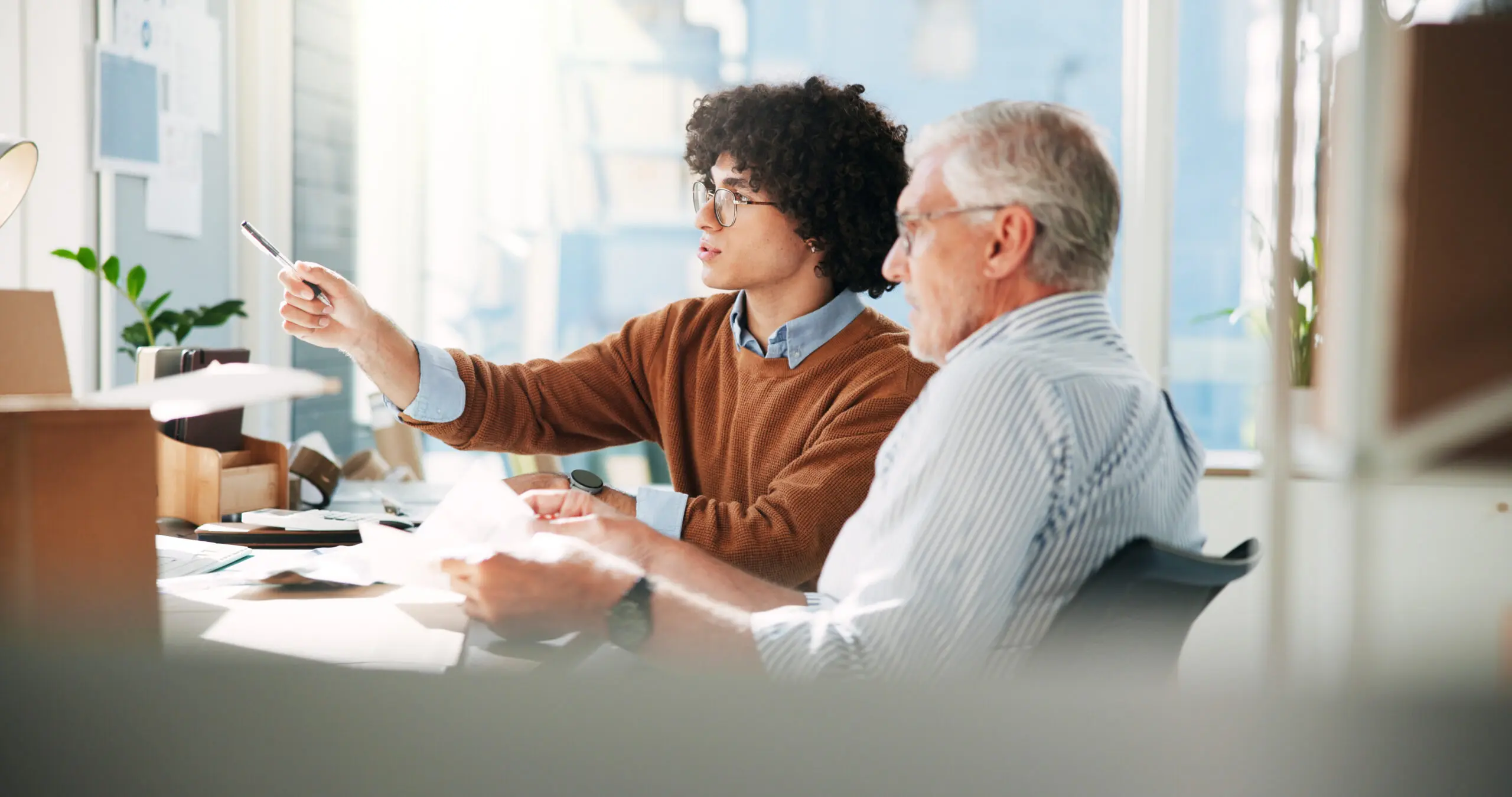Adherent (formerly Compliance & Risks). Two men, one young with curly hair and one older with gray hair, are seated at a desk, both looking and pointing with a pen towards something out of frame.