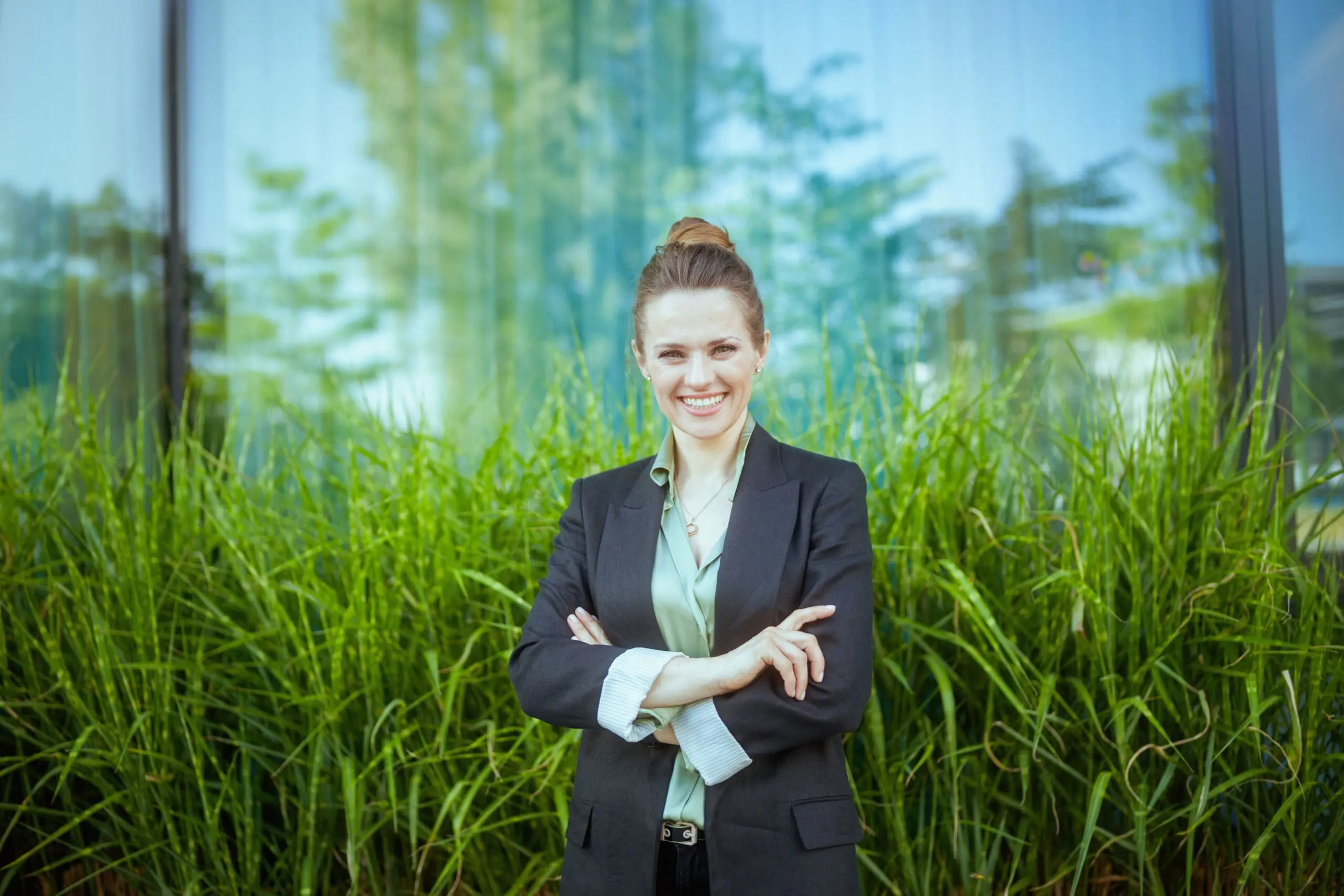 Adherent (formerly Compliance & Risks). A smiling woman in a dark blazer and light green shirt stands with crossed arms in front of green plants and a reflective glass building.