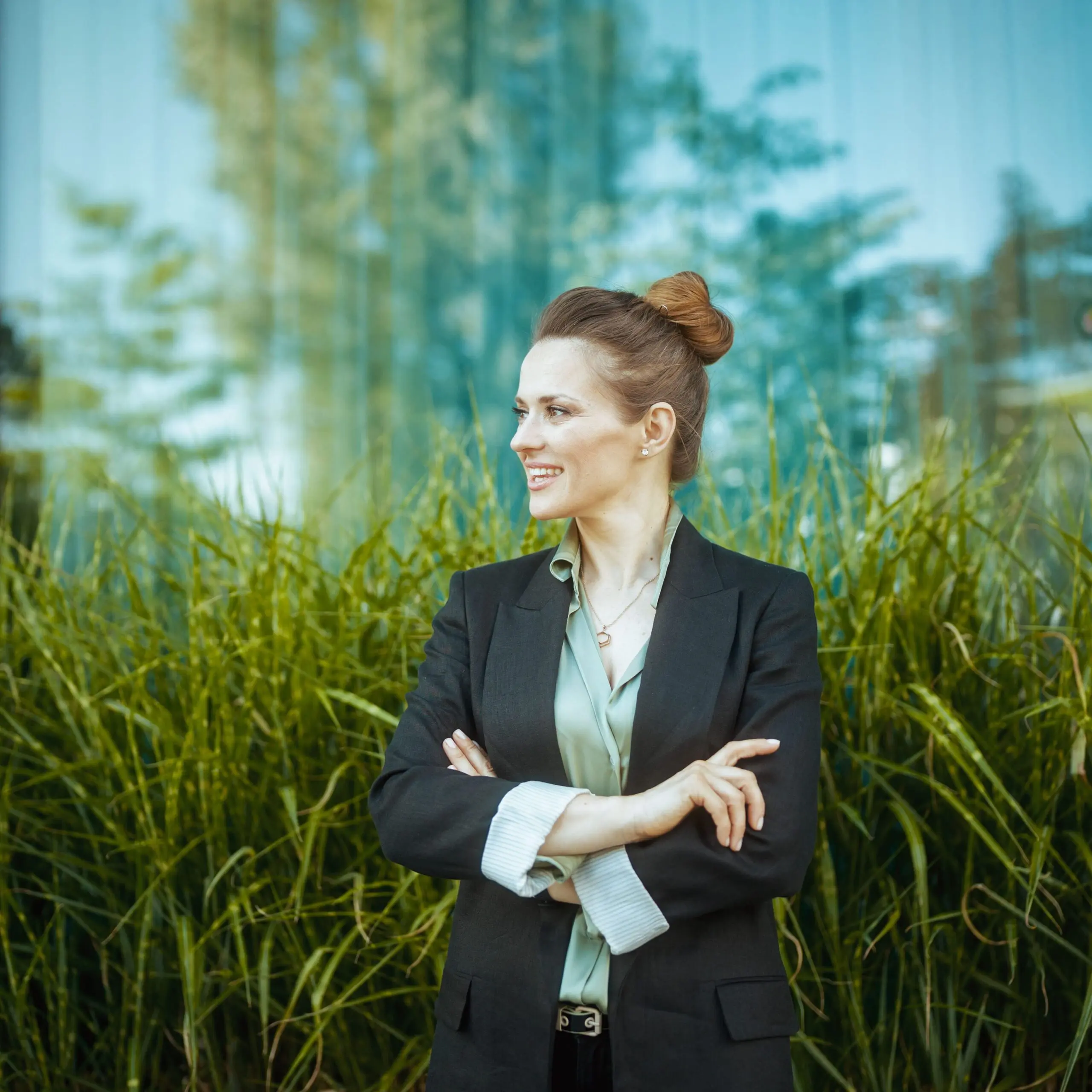 A smiling woman in a black blazer and light green blouse stands with crossed arms in front of green foliage and a reflective glass building.