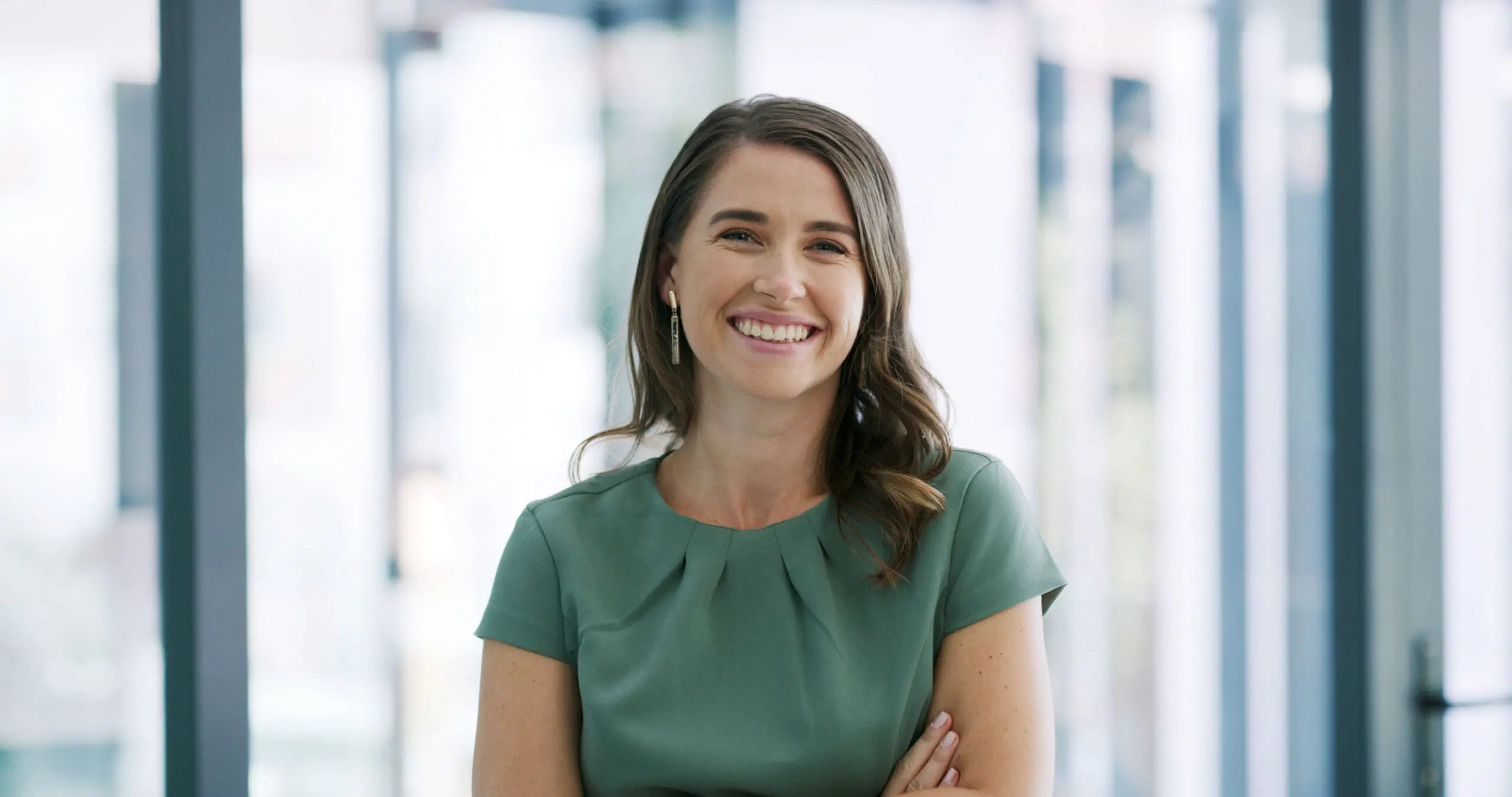 A smiling woman with light brown hair and a green top, arms crossed, looking at the camera in a modern office setting.