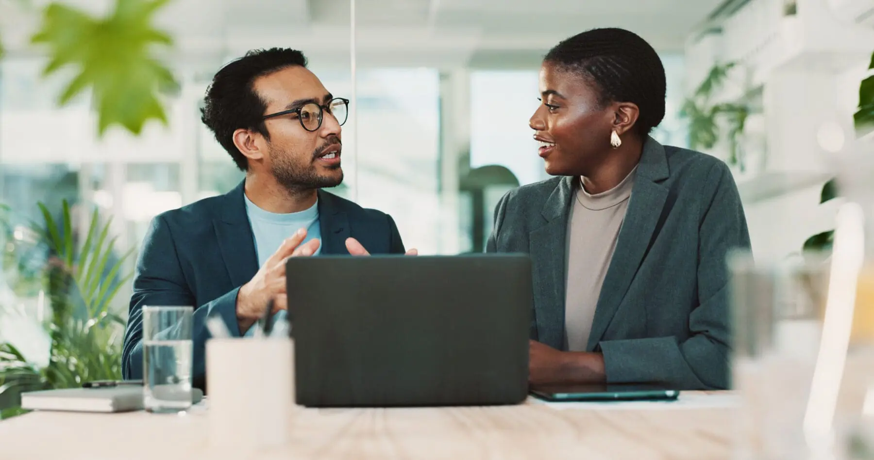 Adherent (formerly Compliance & Risks). A man and a woman in business attire are seated at a table, facing each other and conversing. A laptop is open between them.