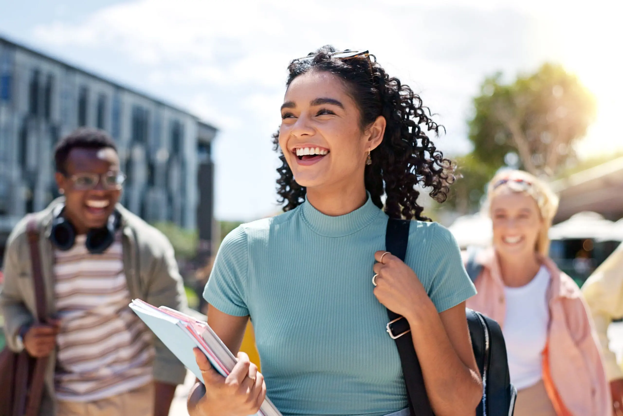 Four diverse students walking outdoors on a sunny day, all smiling. A young woman with curly hair in a teal top is in the foreground, holding books.