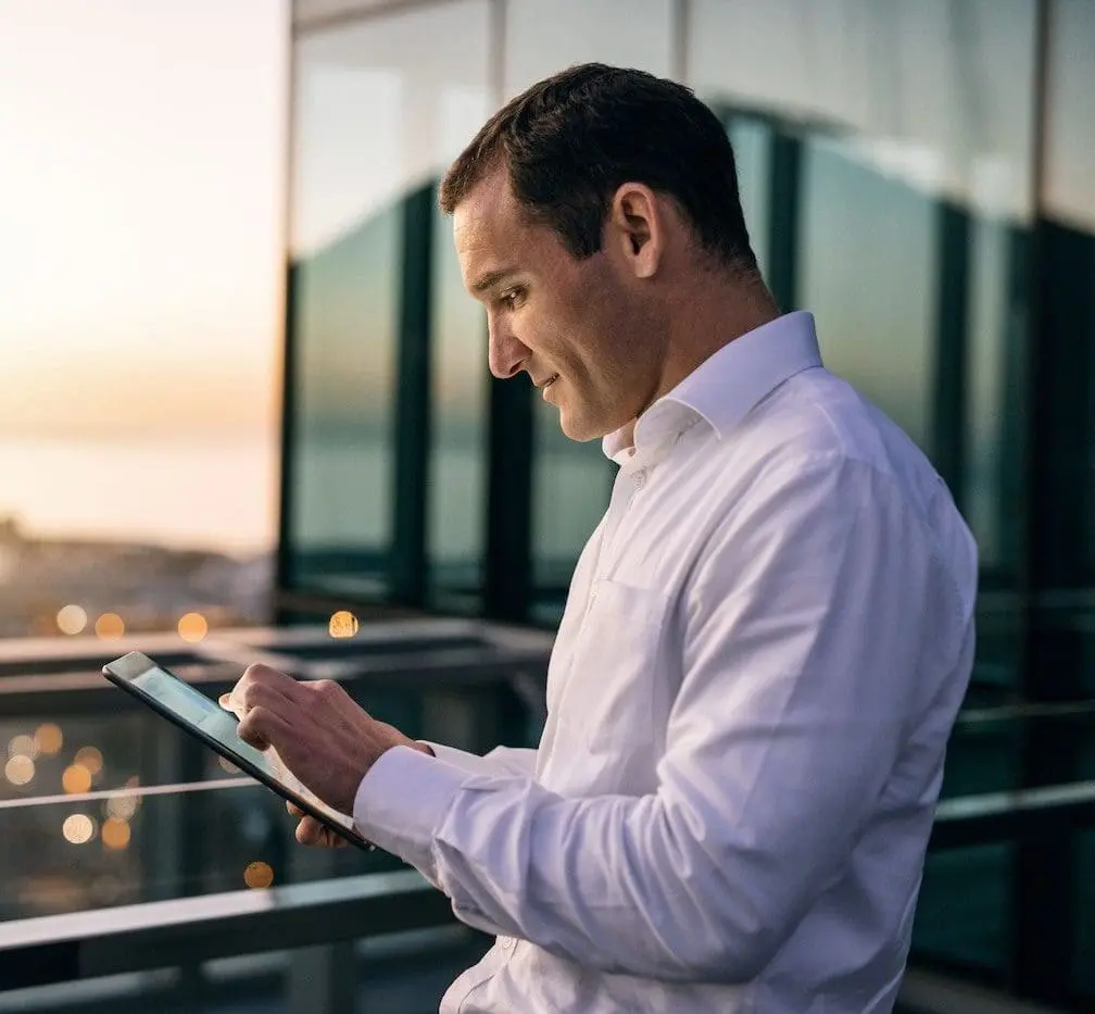 A man in a white shirt uses a tablet on a balcony overlooking a city at sunset.