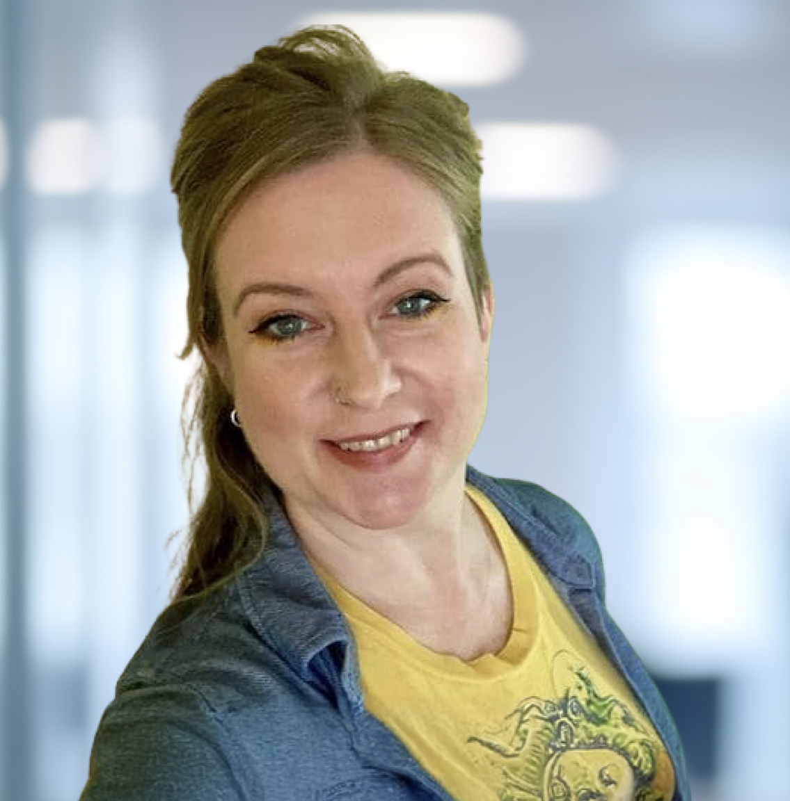 A smiling woman with light brown hair, a nose ring, and a yellow graphic t-shirt under an open blue shirt, looking at the camera.