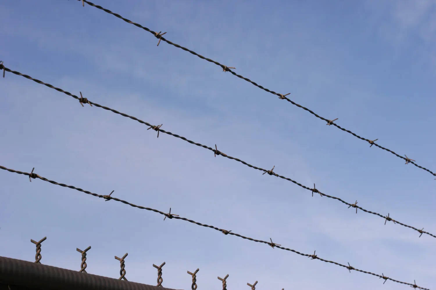 Five strands of barbed wire with sharp barbs against a light blue sky with faint clouds. The top of a dark, corrugated fence with twisted metal spikes is visible at the bottom.