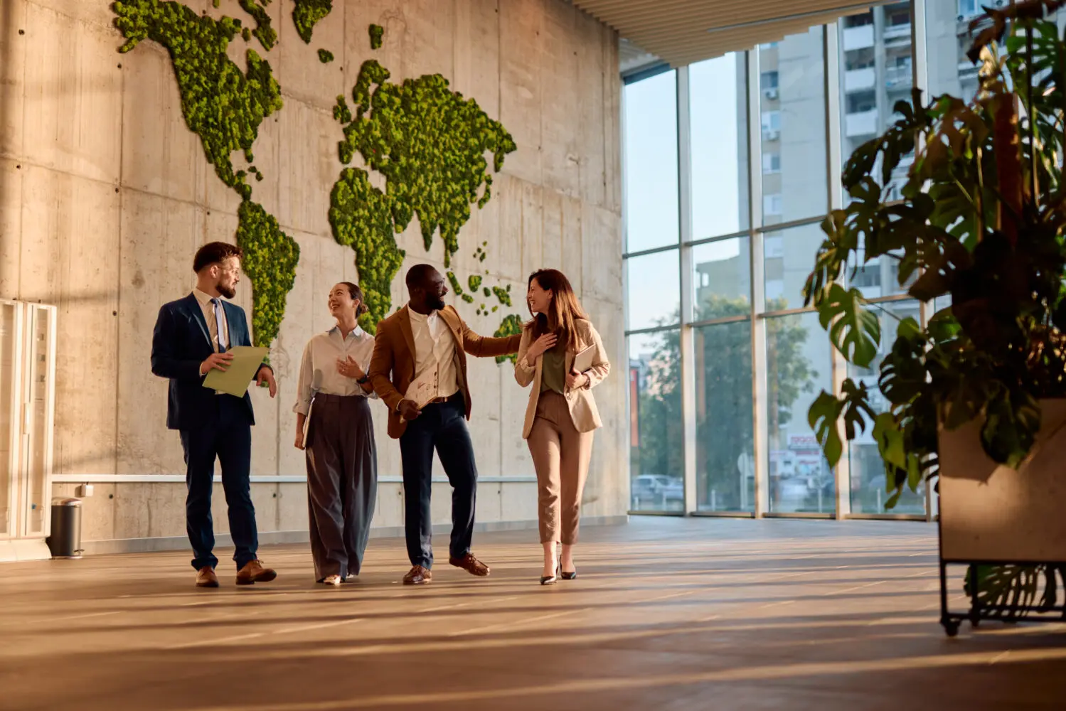 Four diverse professionals, two men and two women, walk and converse in a bright office lobby with a large green moss world map on a concrete wall and large windows.