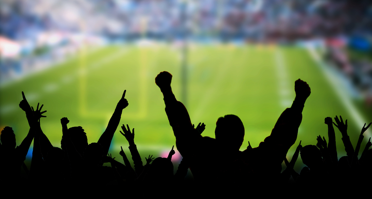 Silhouette of a cheering crowd with raised arms in front of a blurred football stadium.