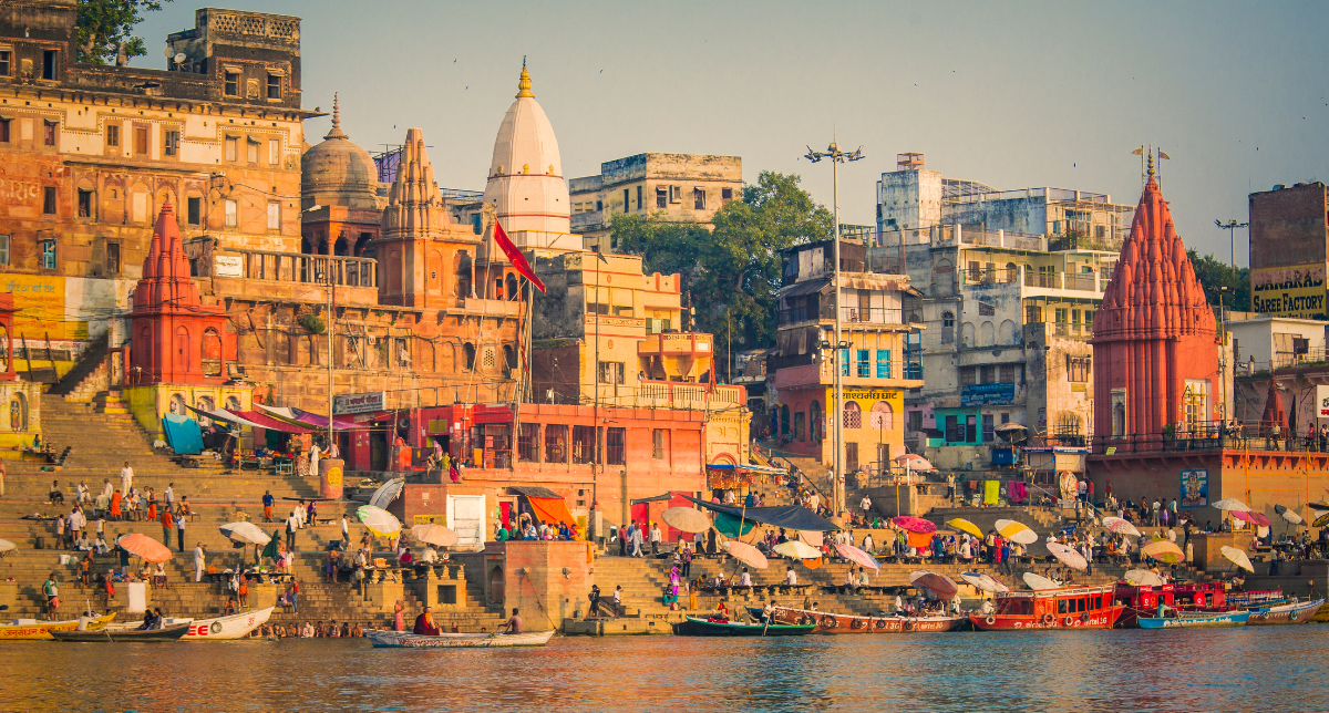 A wide view of a bustling riverbank with stone steps (ghats) leading to water. Numerous people are on the ghats and in boats. Multi-colored buildings, including temples with spires and domes, line the bank under a bright sky.