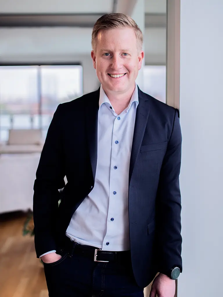 A smiling man in a dark blue suit jacket, light blue shirt, and dark jeans, leaning against a wall.