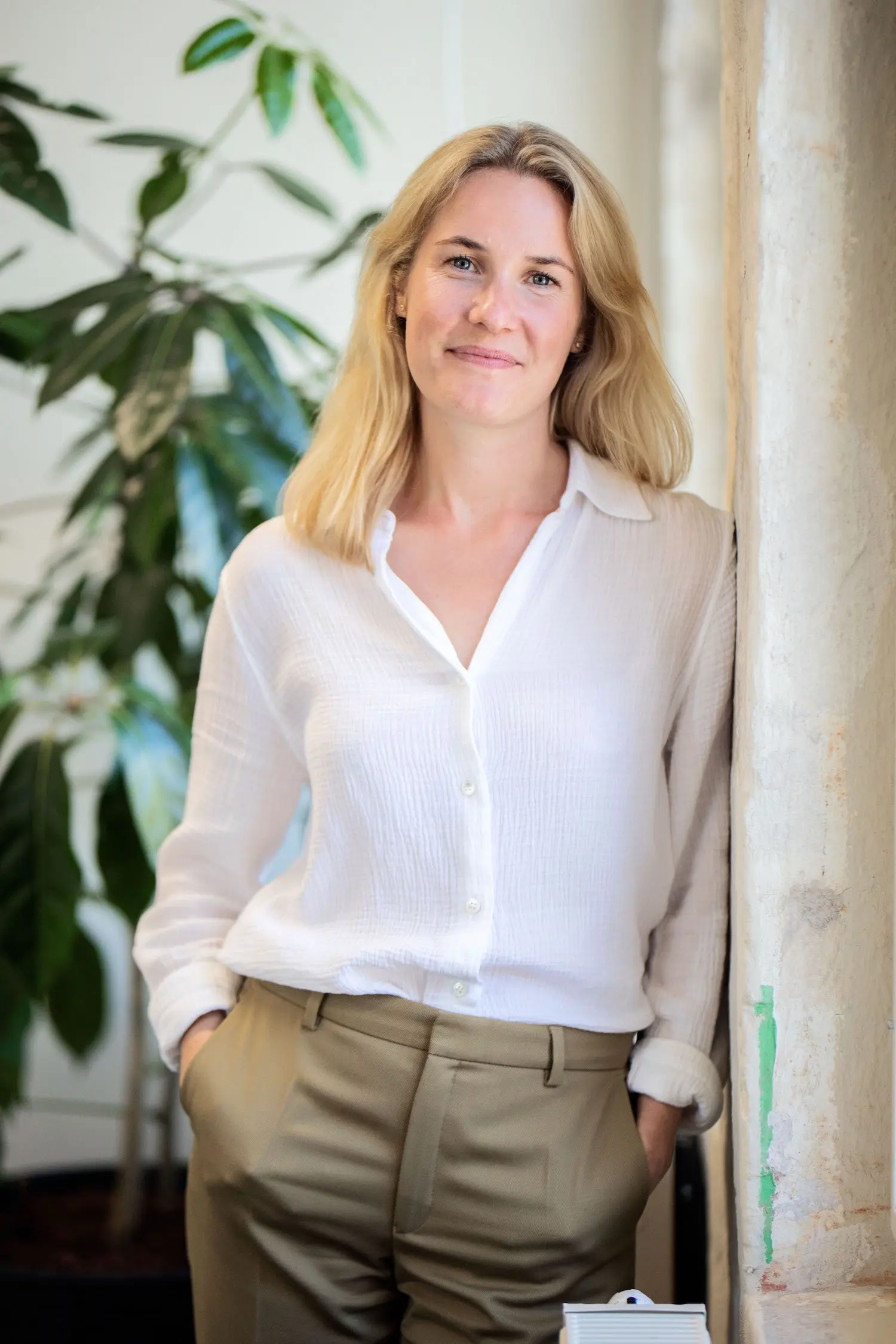 A smiling woman with blonde hair, wearing a white shirt and khaki trousers, stands leaning against a light-colored pillar.