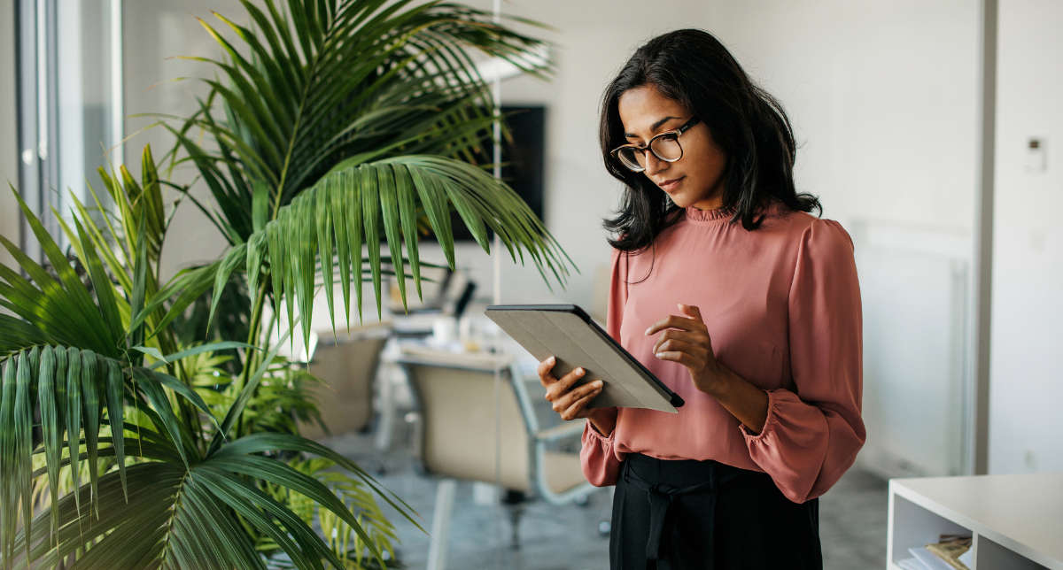 A woman in a pink blouse and glasses uses a tablet in a bright office next to a large green plant.