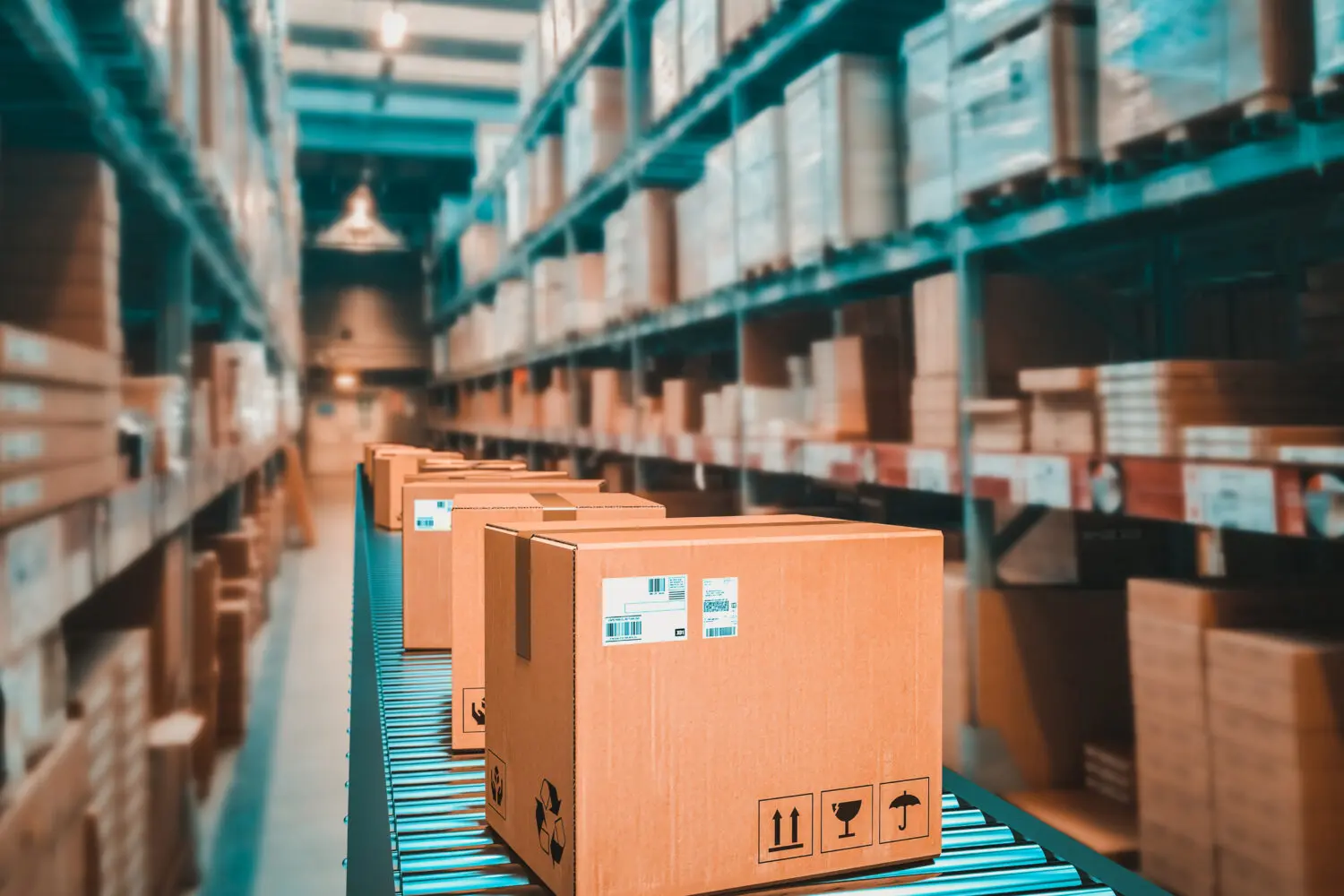 Adherent (formerly Compliance & Risks). Cardboard boxes on a blue roller conveyor belt in a large, blurred warehouse with tall shelves filled with inventory.
