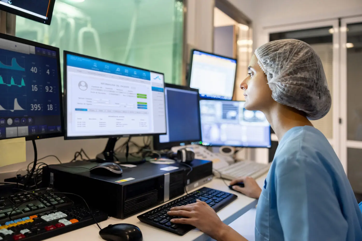 Side view of a medical professional in scrubs and a hair net, focused on multiple computer screens displaying patient information and medical data.