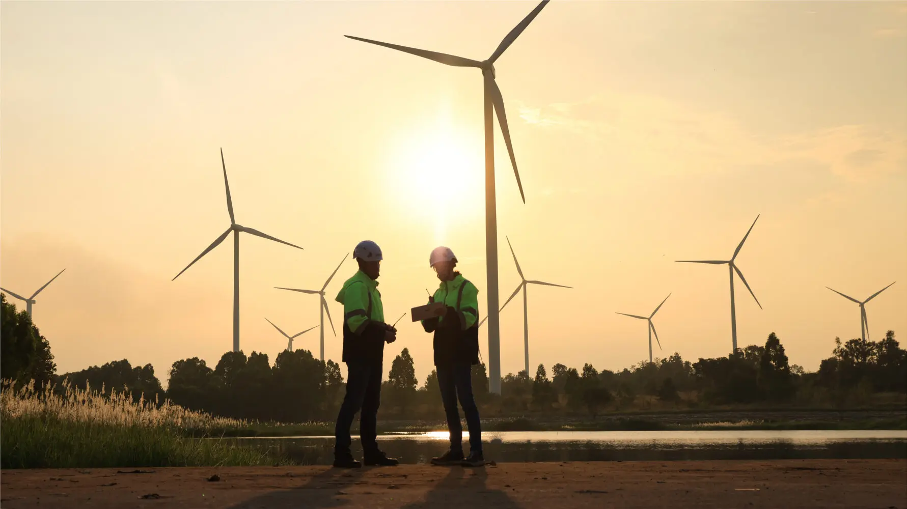 Two silhouetted engineers in hard hats and high-visibility jackets stand on a dirt path, discussing operations at a wind farm during sunset, with multiple large wind turbines in the background.
