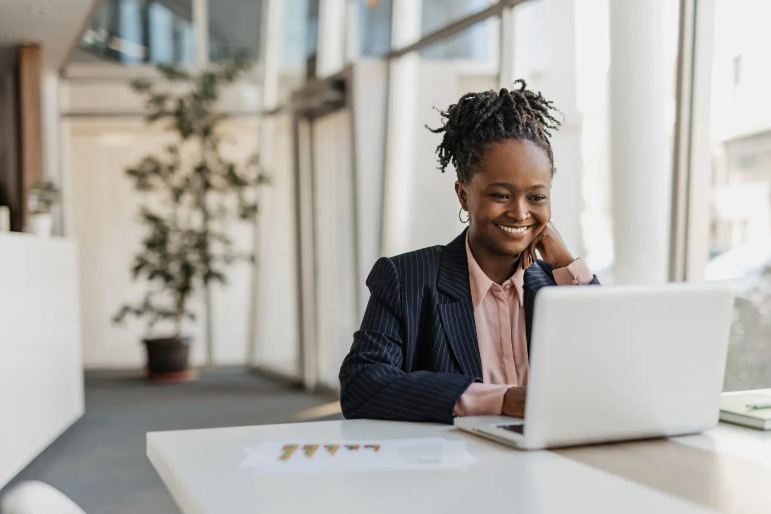 A smiling woman works on a laptop at a white table in a bright office.
