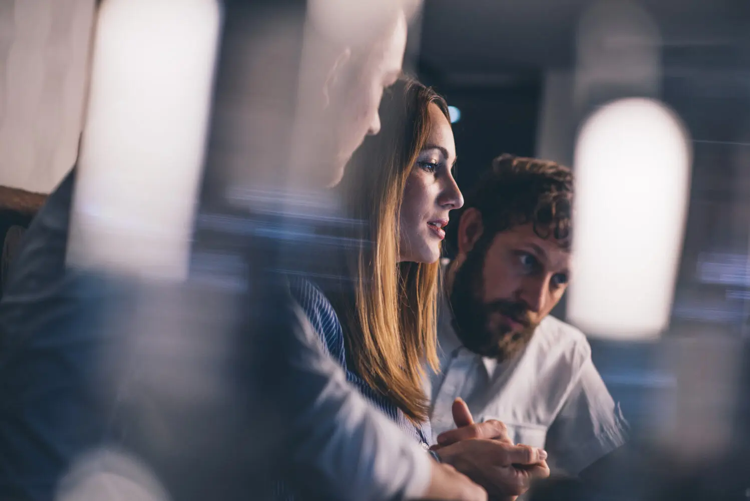 Three people in a meeting, with a woman in profile at the center, a bearded man to her right, and another person partially visible. Light reflections are prominent in the foreground.