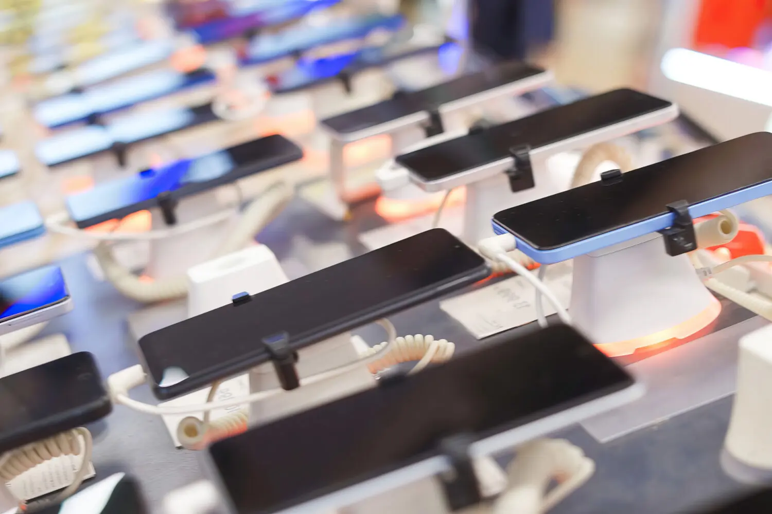 Multiple smartphones on display stands with security cables in an electronics store, some stands illuminated with orange light.