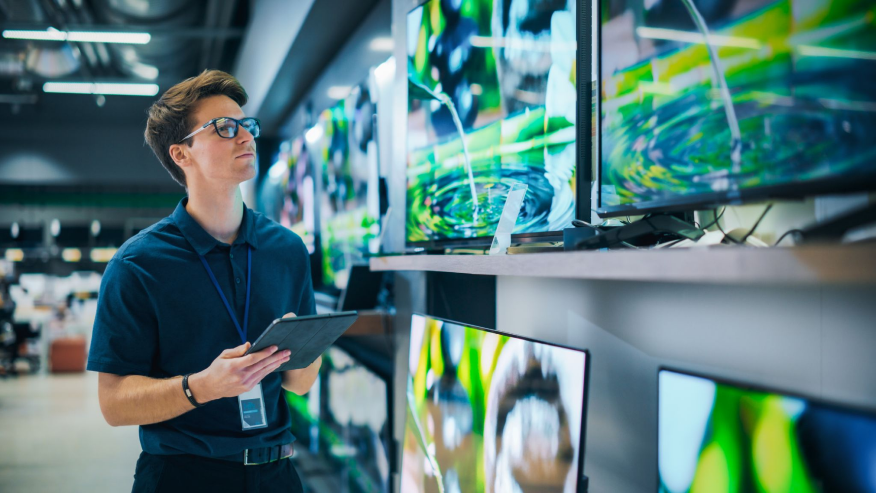 A man in glasses and a blue polo shirt holds a tablet, looking up at multiple flat-screen TVs displaying colorful images in an electronics store.
