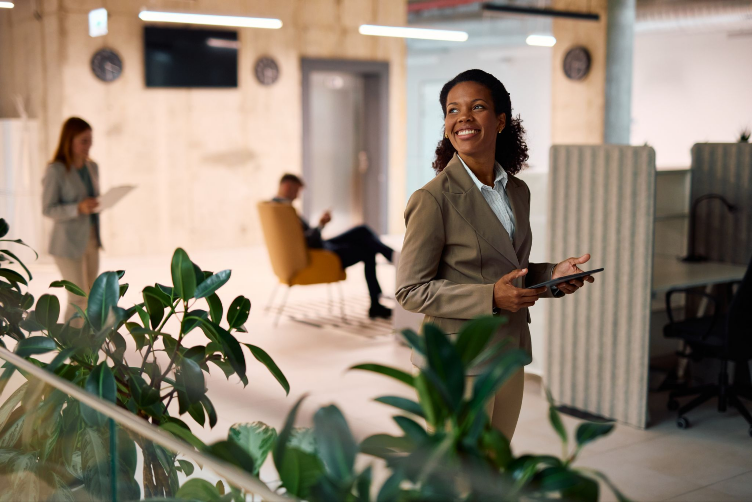 A smiling Black woman in a tan suit holds a tablet in an office, with blurred colleagues and plants in the background.