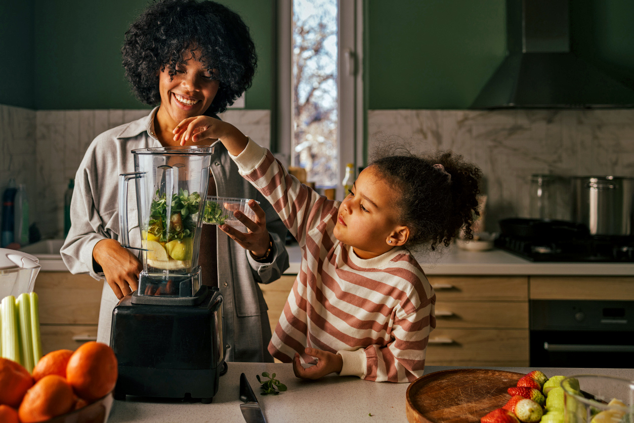 A smiling woman and a young girl in a kitchen, adding green ingredients to a blender to make a smoothie. Oranges and celery are on the counter.