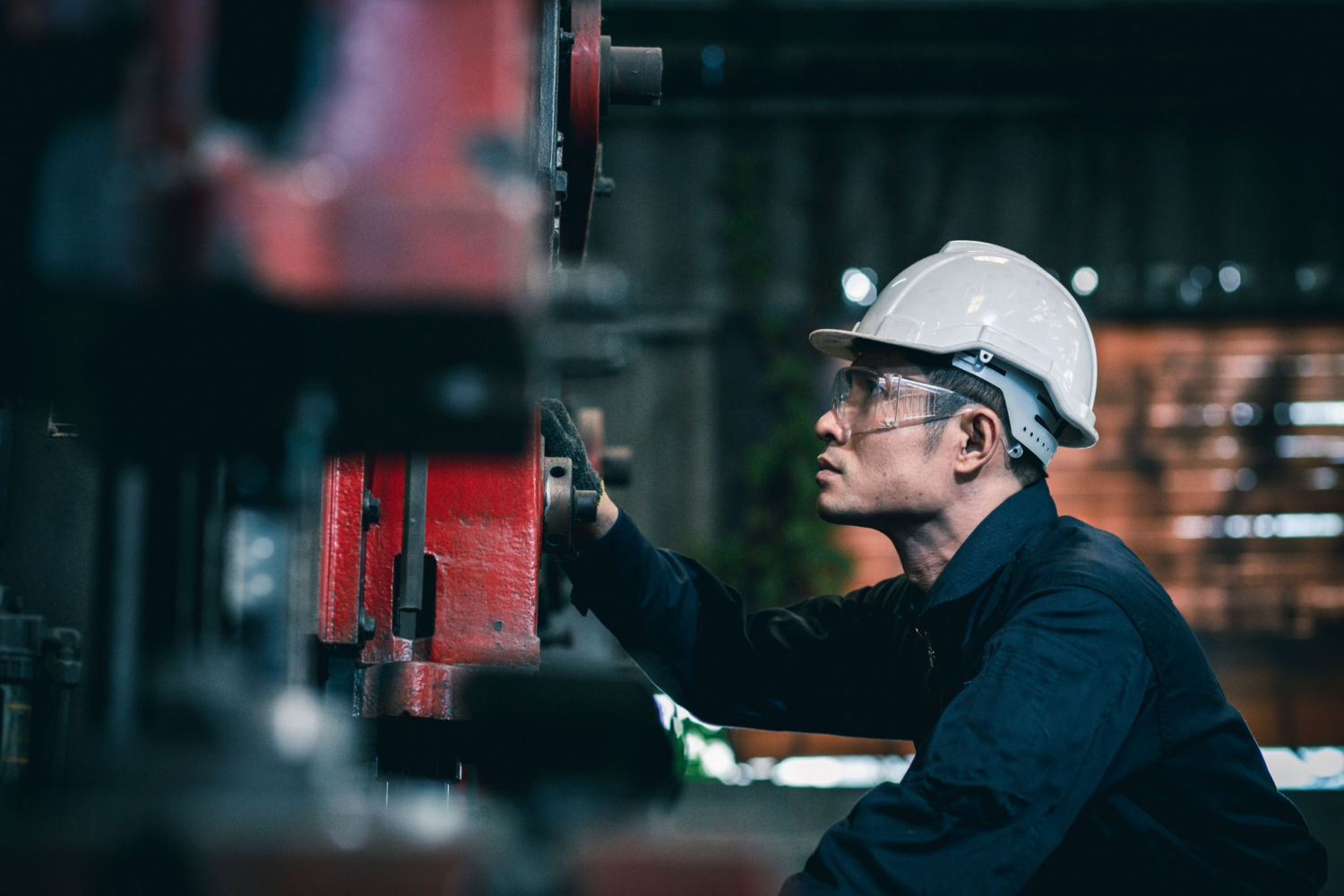 Profile view of a man in a white hard hat and safety glasses, inspecting a large red industrial machine with his gloved hand.