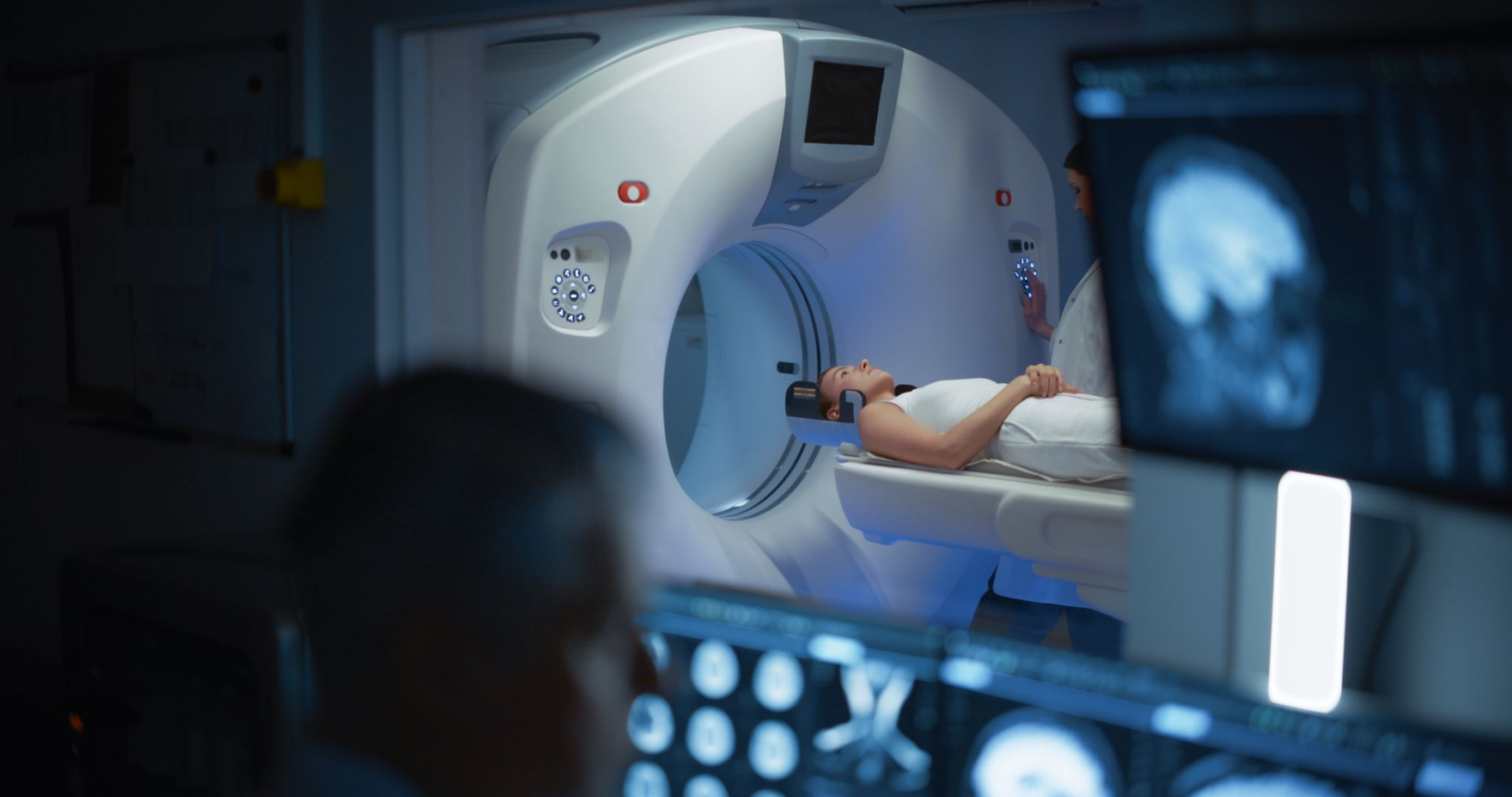 A patient lies on a medical scanner bed, entering a large white CT or MRI machine with blue light. A medical professional stands next to the scanner, and in the foreground, monitors display medical scans.
