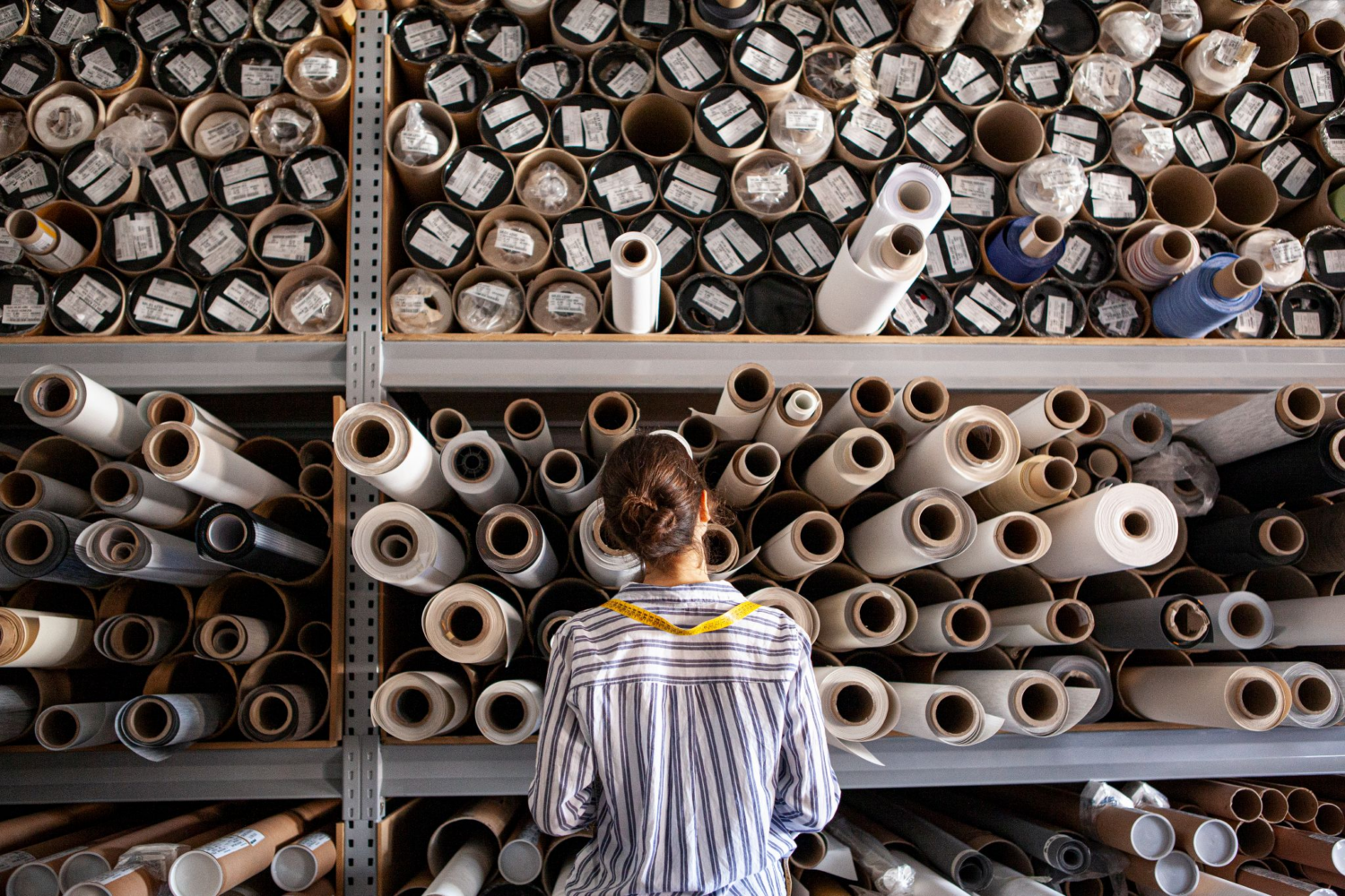 Rear view of a woman with a measuring tape around her neck, standing in front of tall shelves filled with numerous rolls of fabric.