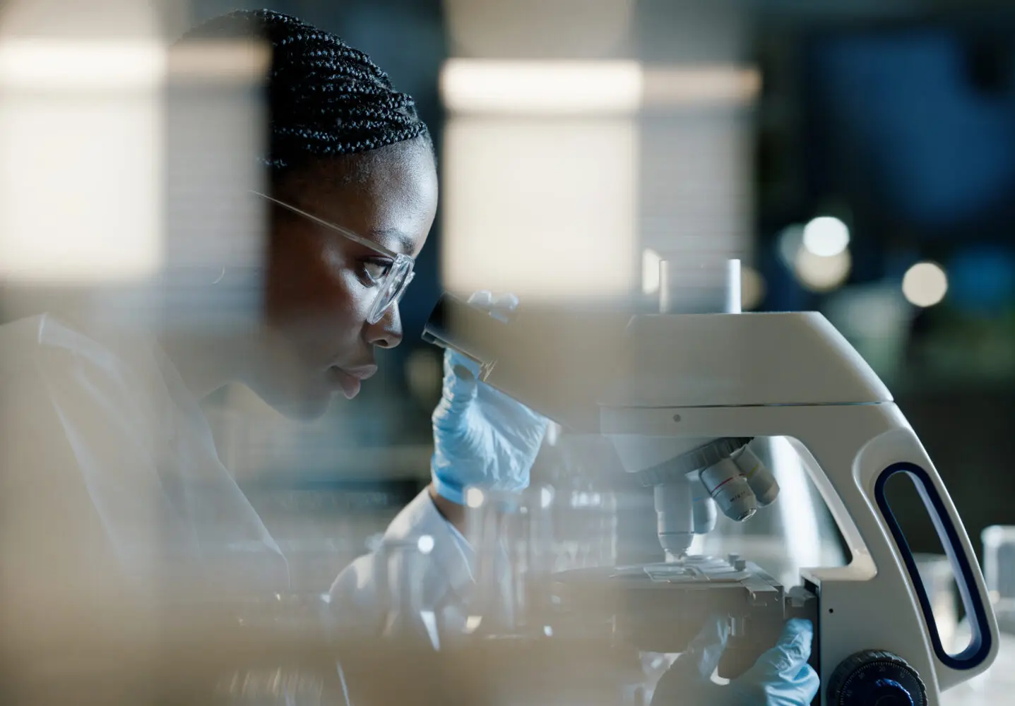 Side profile of an African American woman in a lab coat and gloves, looking through a microscope.