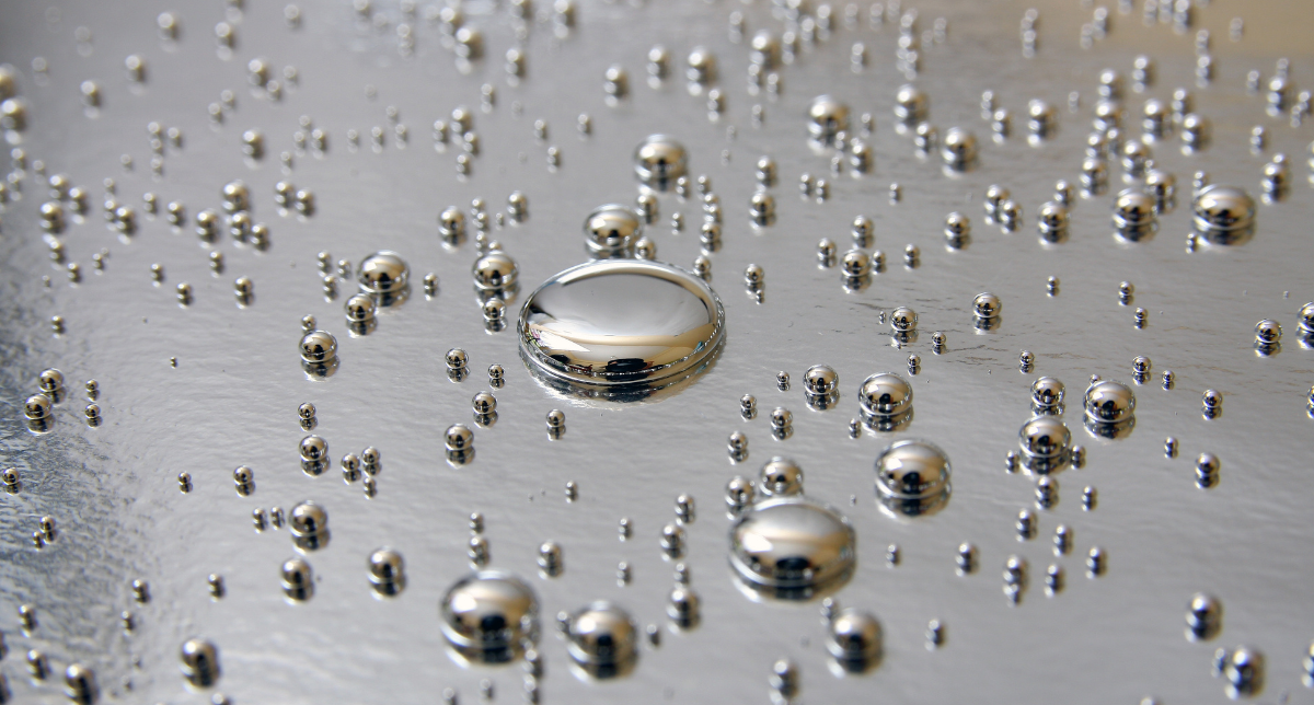 Numerous silver, spherical liquid metal droplets of various sizes scattered on a reflective gray surface.