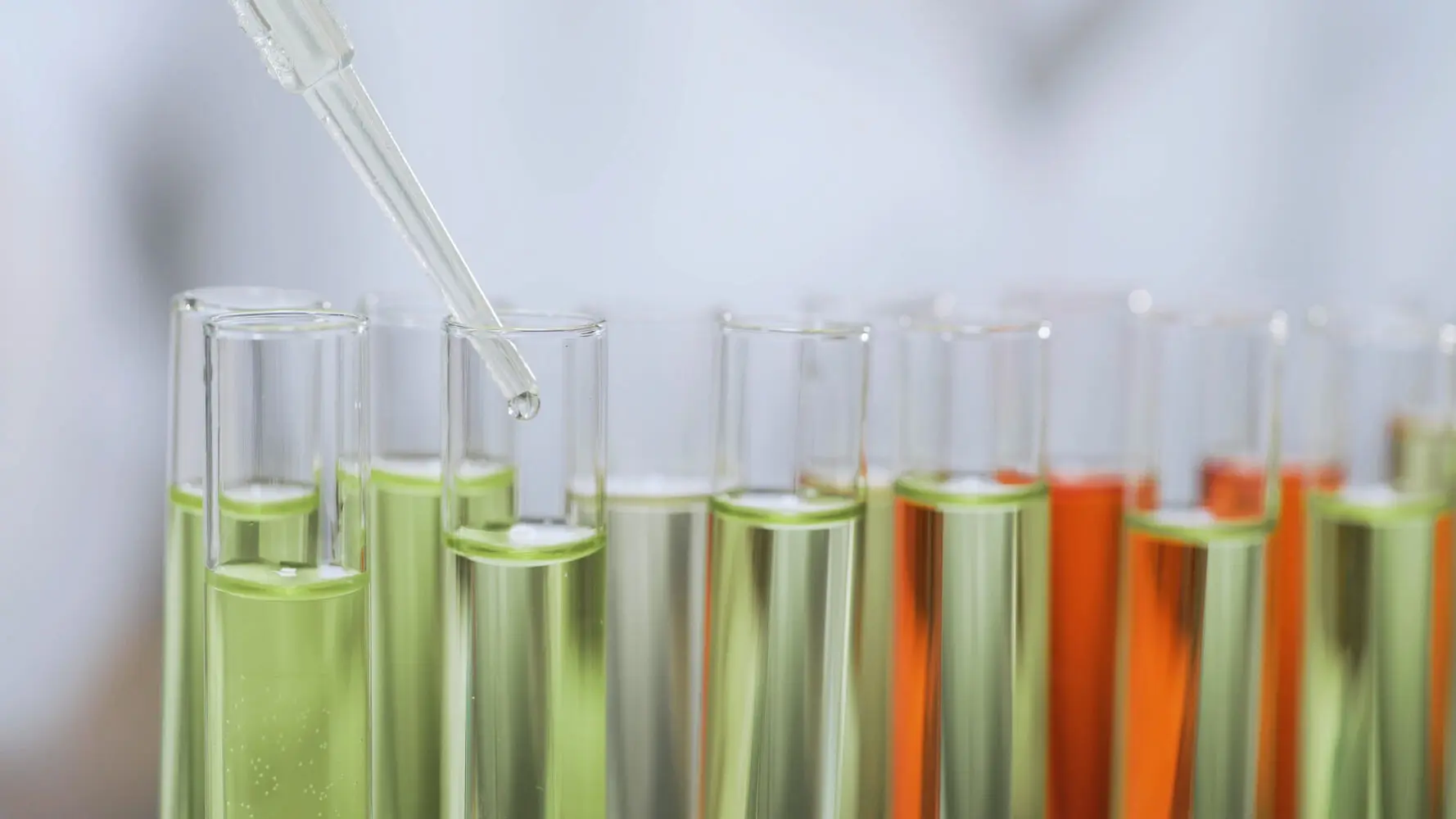 A close-up of a glass dropper adding a clear liquid drop into a test tube filled with green liquid, surrounded by other test tubes containing green and orange liquids.