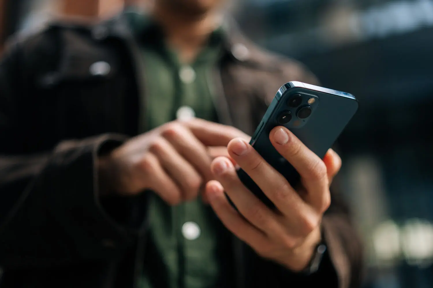 Close-up of hands holding a dark blue smartphone with a triple camera system, with a finger poised to touch the screen. The background is blurred.