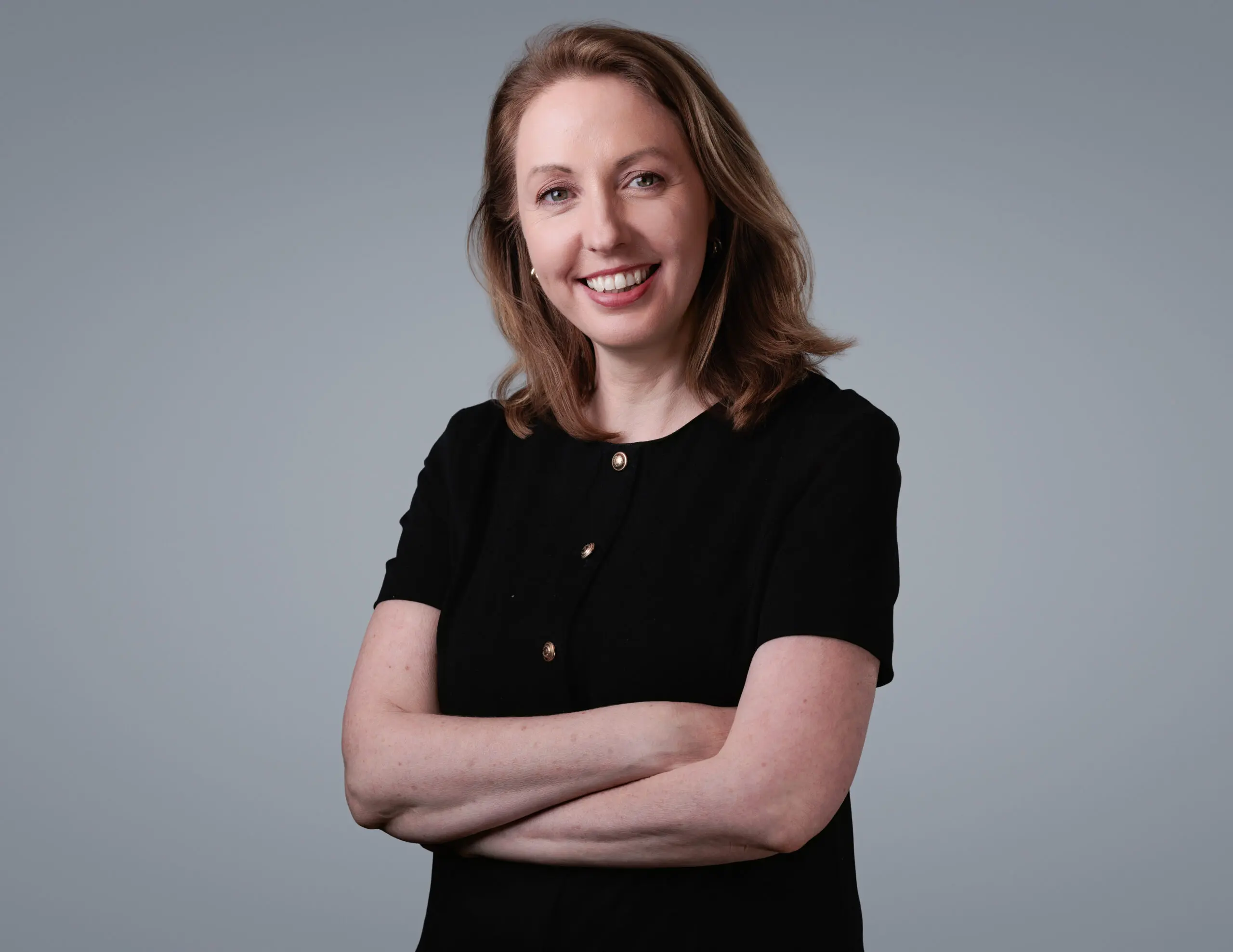 Headshot of a smiling woman with light brown hair, wearing a black top with gold buttons, arms crossed, against a gray background.