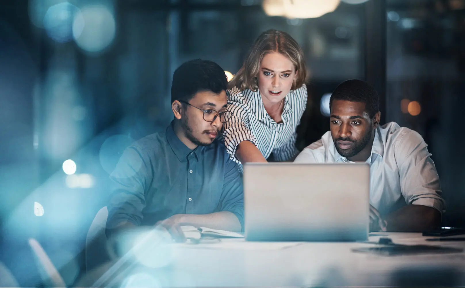 Three diverse colleagues, two men and one woman, intently looking at a laptop screen in a dimly lit office.