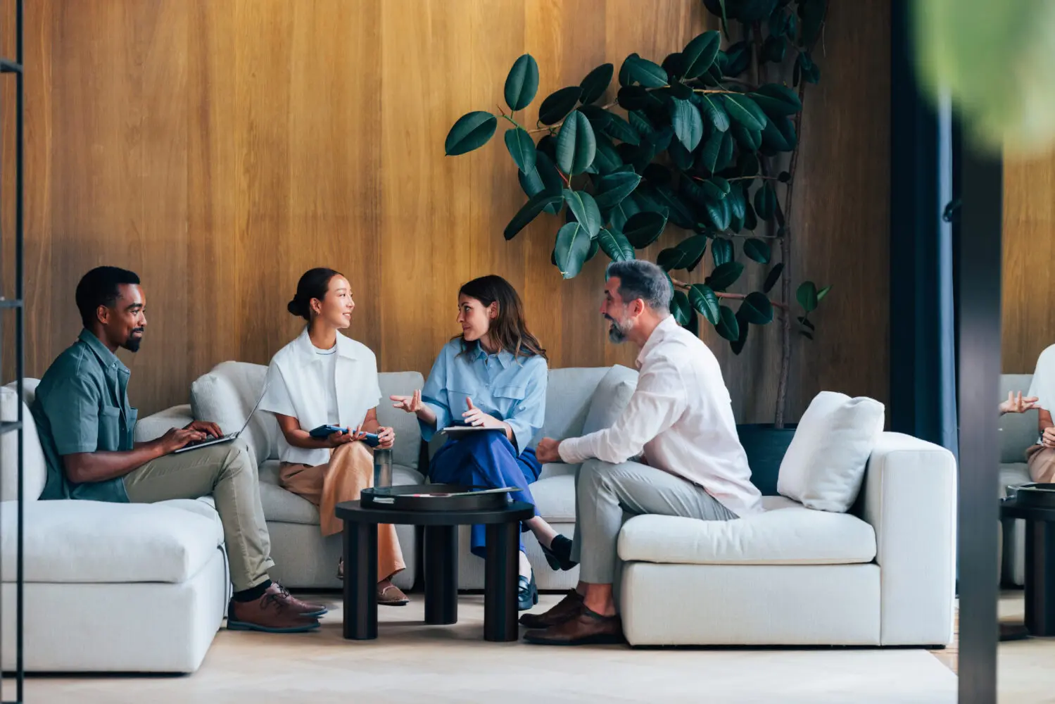 Four professionals, two men and two women, seated on light-colored sofas in a modern office lounge, engaged in a discussion. One man is typing on a laptop, and one woman is gesturing while speaking.