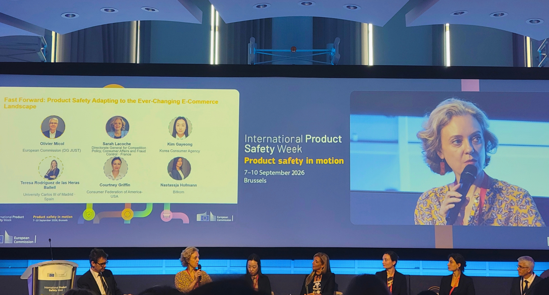 A conference panel discussion with six individuals seated below a large screen. The screen displays the event title 'International Product Safety Week,' 'Product safety in motion,' and '7-10 September 2026, Brussels.' It also shows a presentation on 'Product Safety Adapting to the Ever-Changing E-Commerce Landscape' with headshots and names of six panelists. A close-up of a woman speaking into a microphone is prominently featured on the right side of the screen.