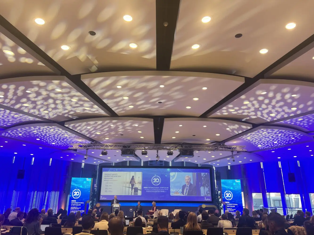 A conference room with a speaker on stage, a large screen displaying 'Product Safety Works The Same Way' and '2026 International Symposium', and an audience seated in rows.